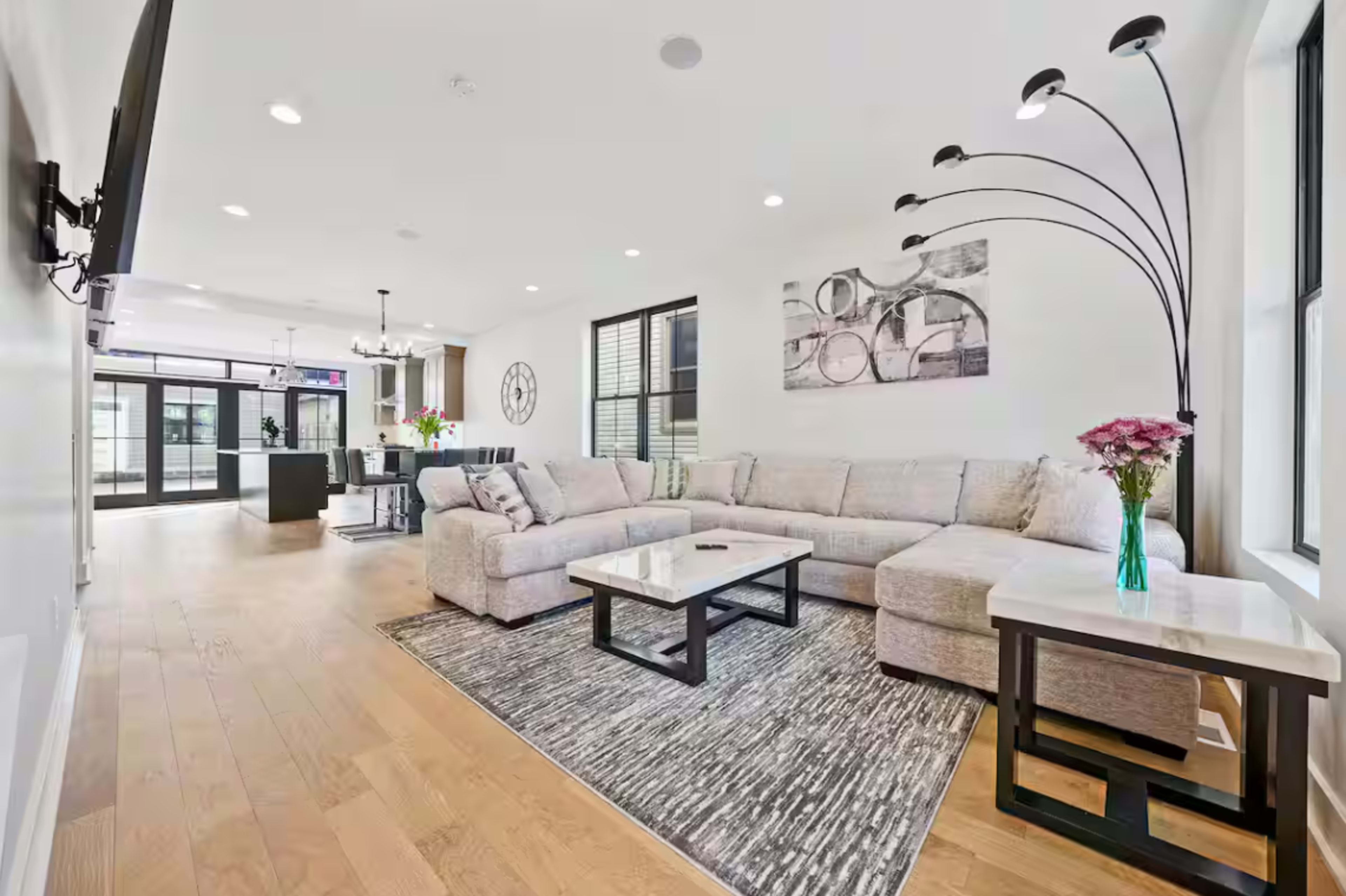 The living room features a light-colored sectional sofa, a coffee table, and a rug, with a large window and an open layout leading to a kitchen area.