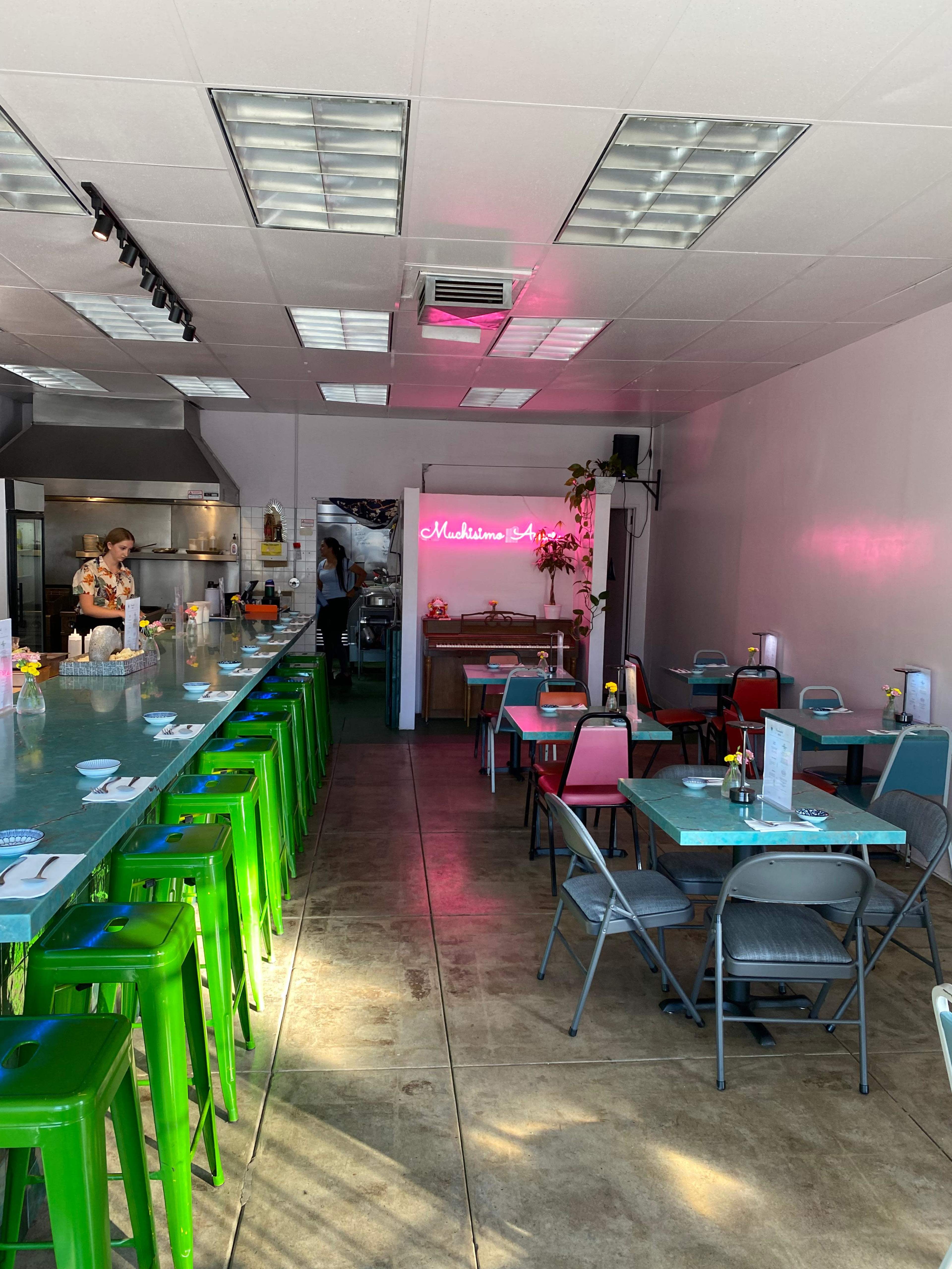 The image shows a brightly lit restaurant interior with colorful seating, including green barstools and blue-gray tables, and a neon sign in the background.