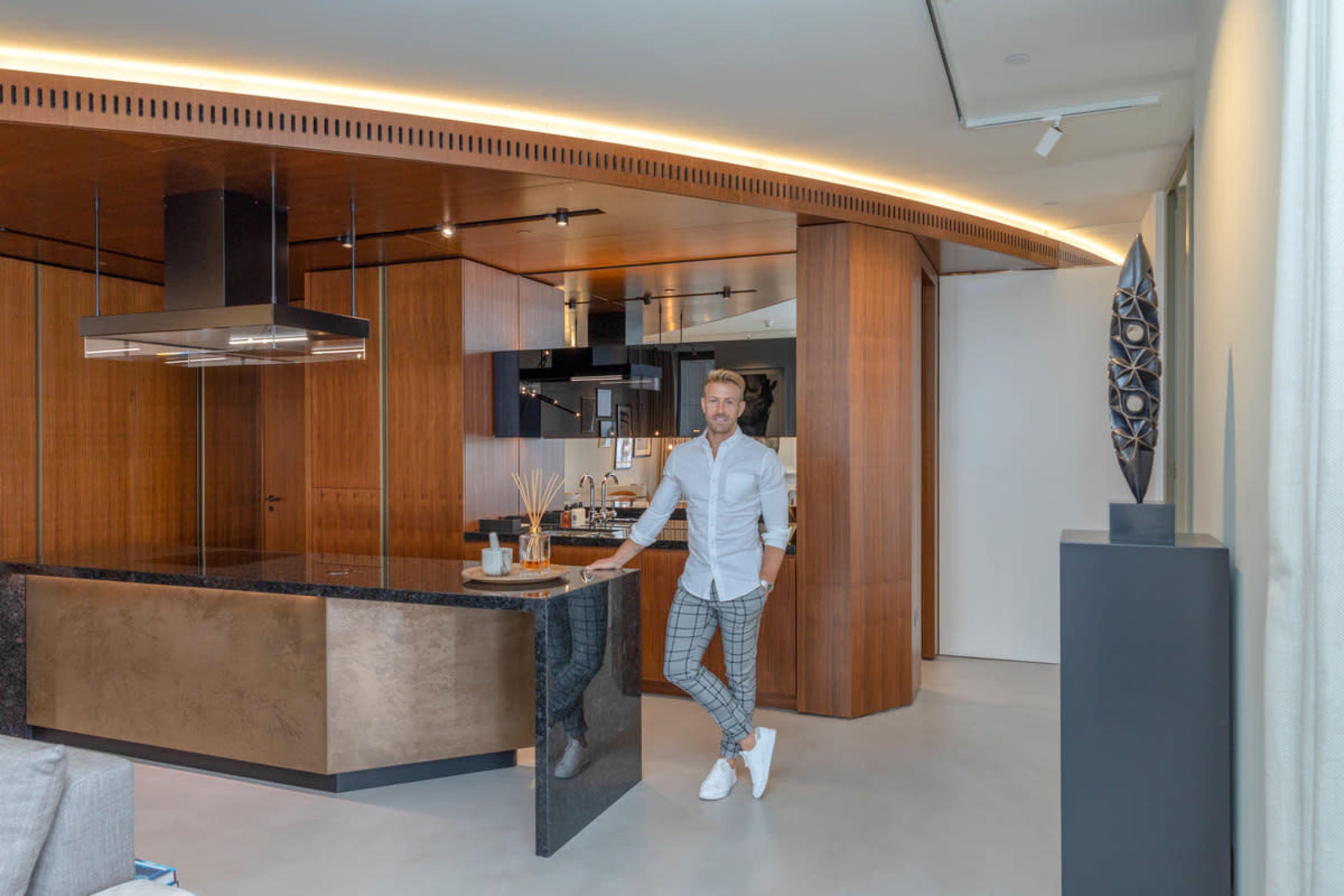 A man stands beside a kitchen island in a modern kitchen featuring wood cabinetry and sleek appliances.