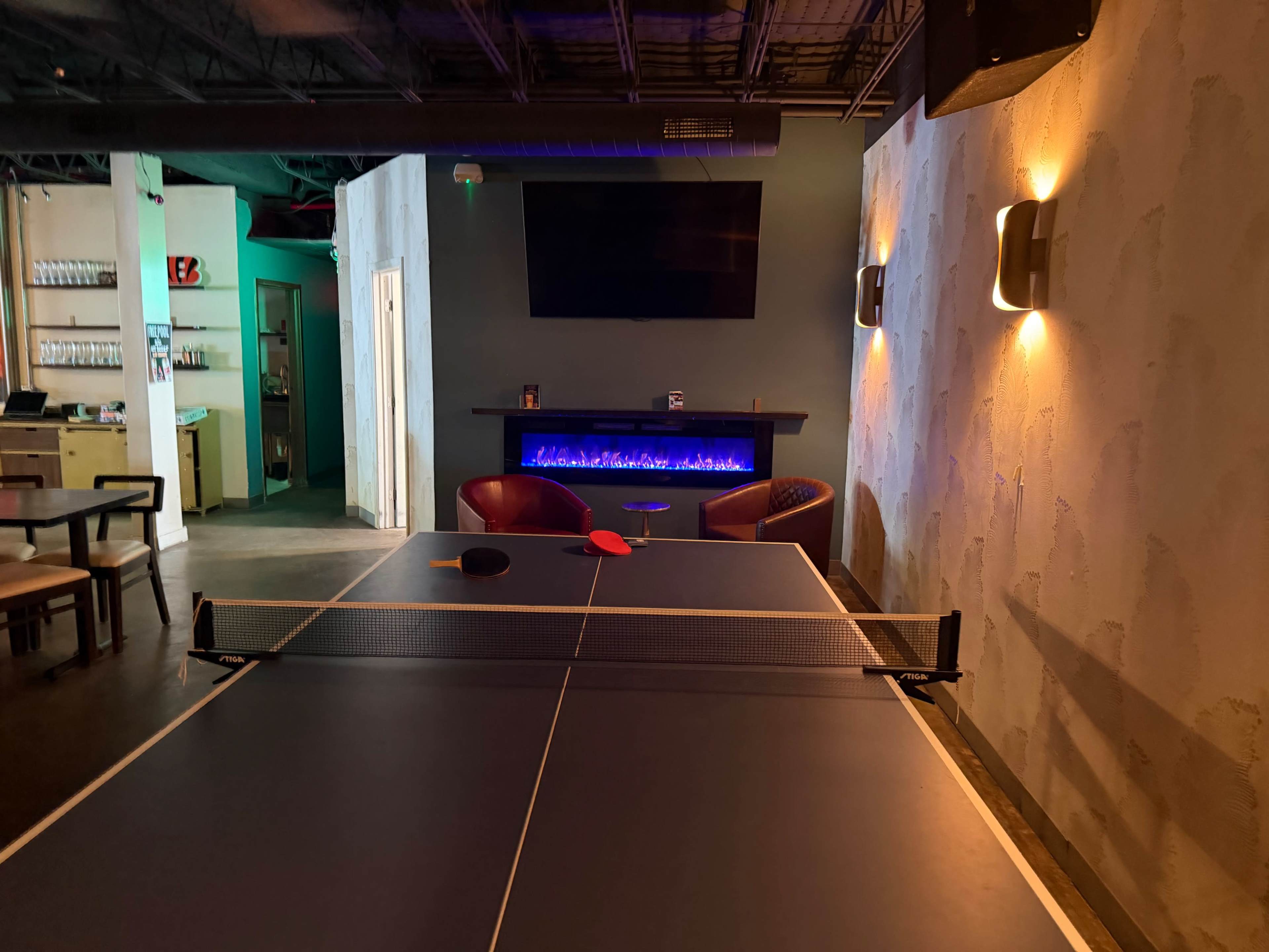 The image shows a ping pong table positioned in a room with a modern aesthetic, featuring a glowing blue electric fireplace and a large flat-screen TV mounted on the wall.