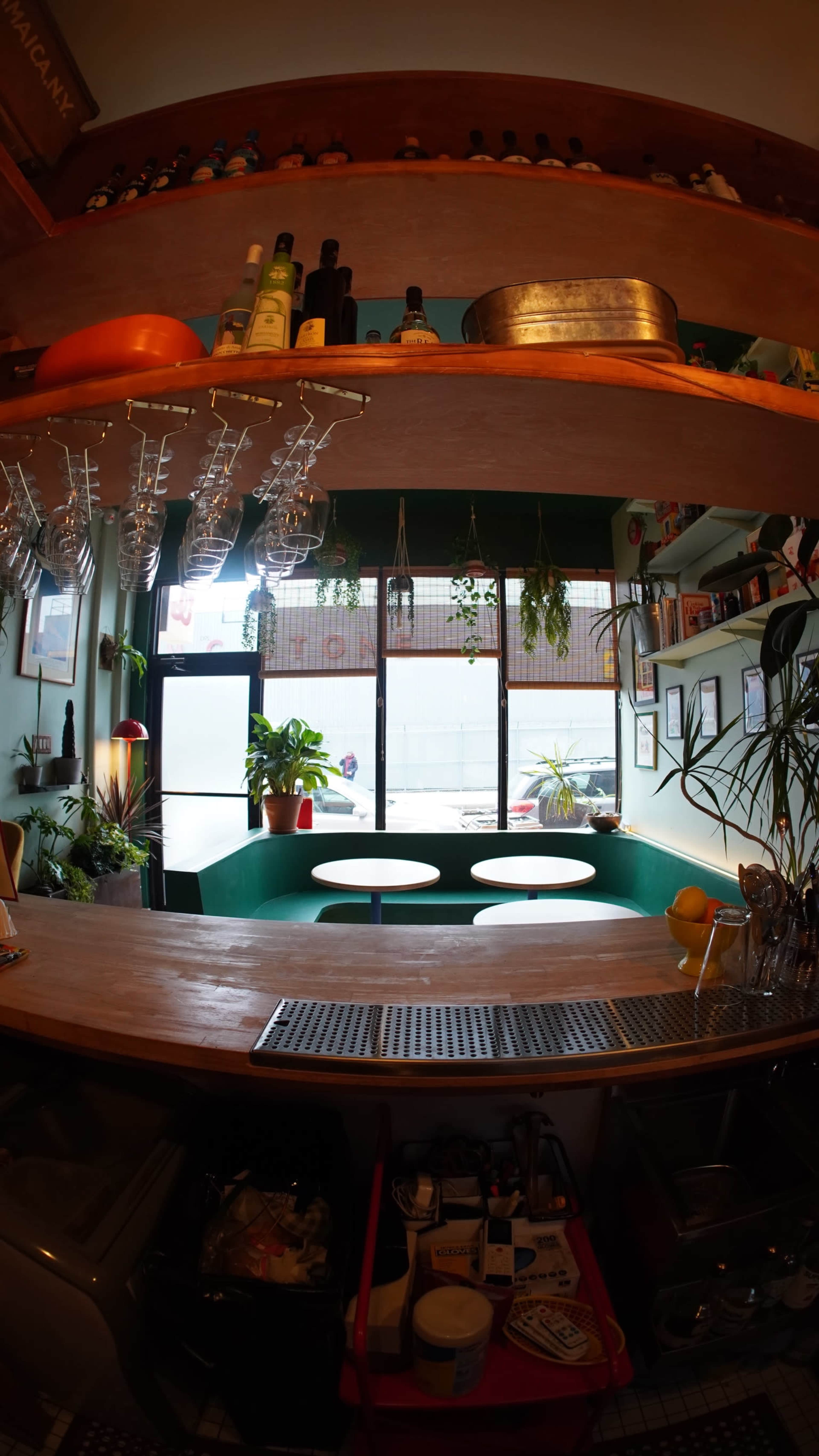 A bar counter with shelves holding various bottles is visible, while green seating and plants are arranged in front of large windows.
