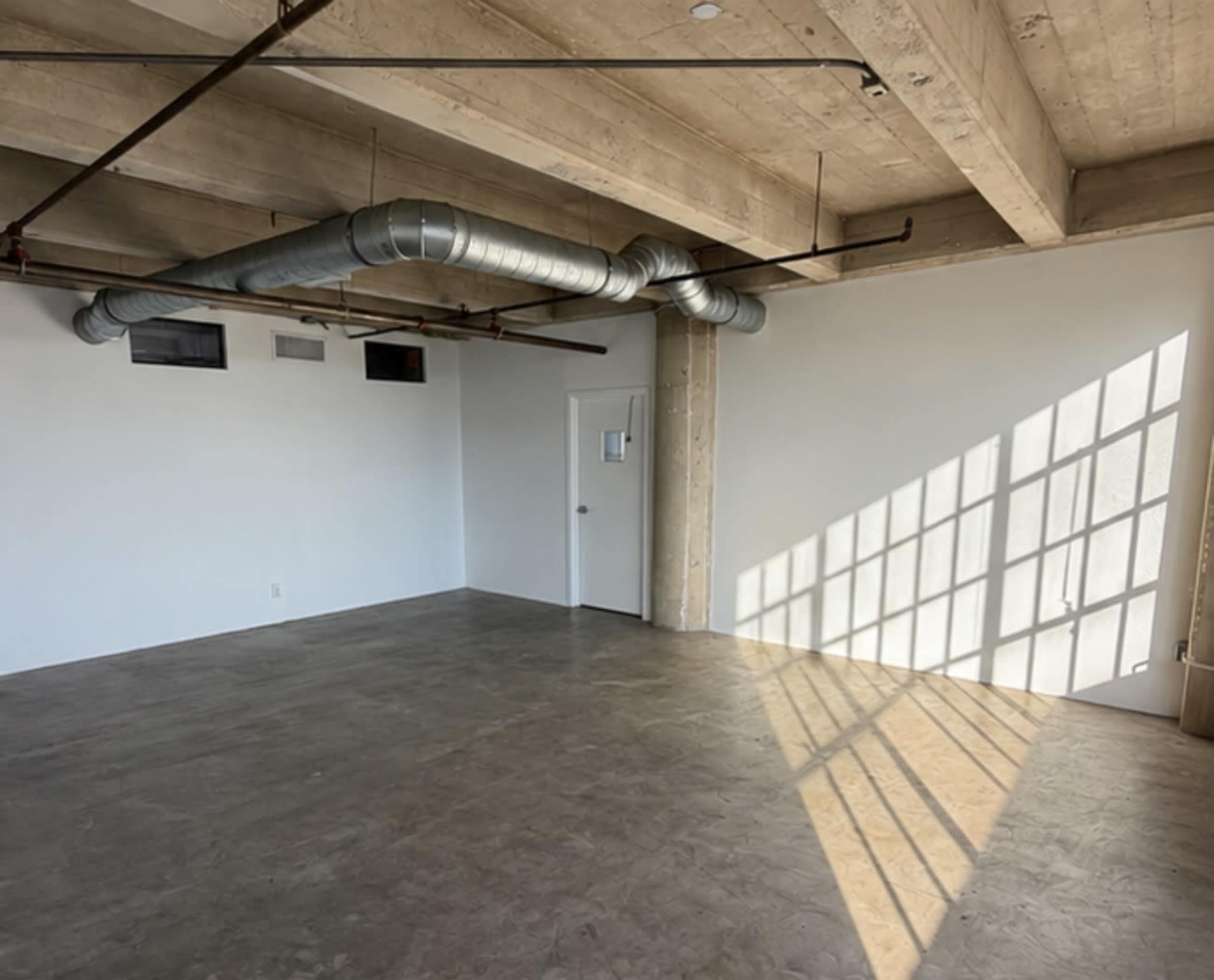 The image shows an empty room with a concrete floor, exposed beams, and shadows cast by sunlight through a window.