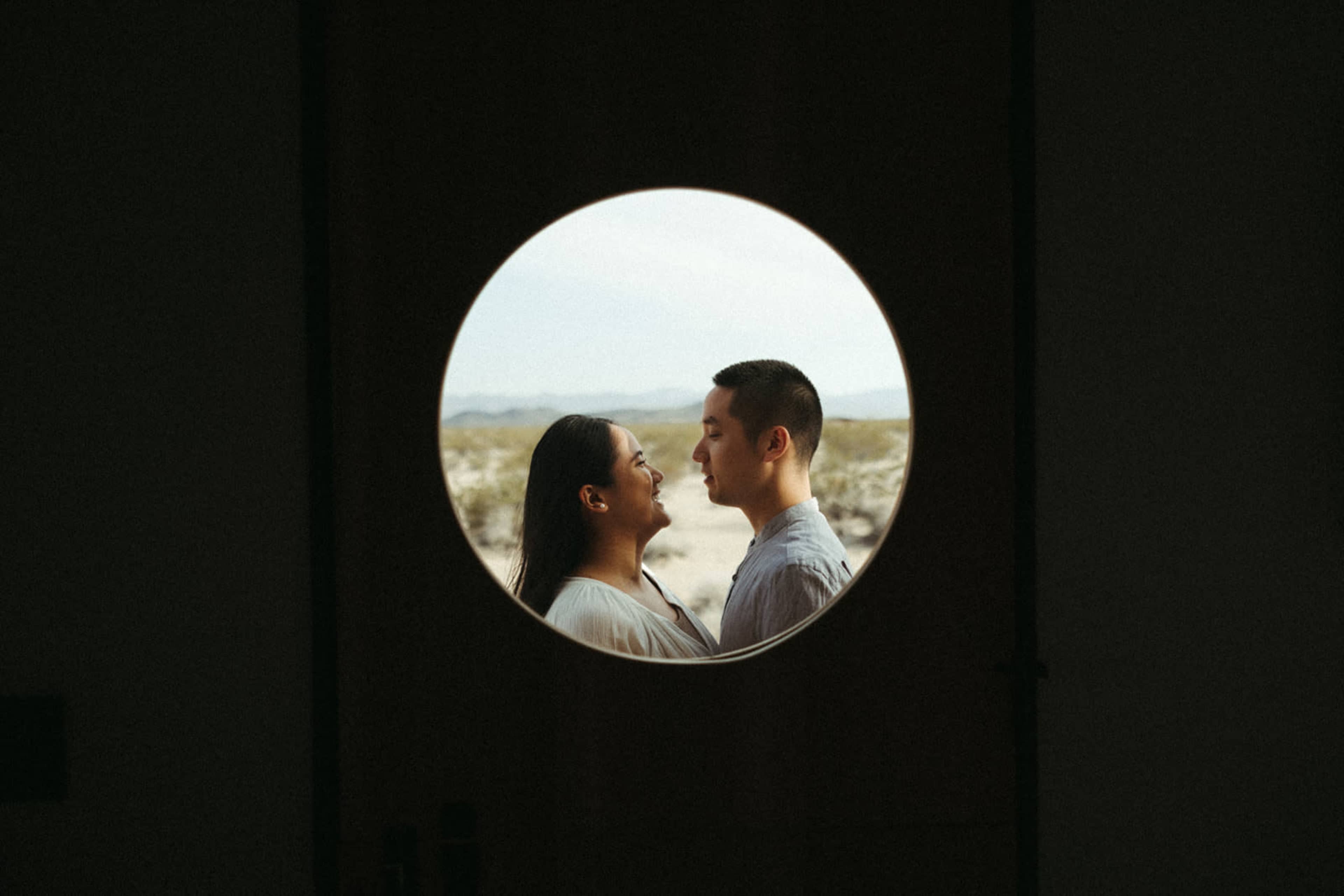 A couple faces each other framed by a circular opening with a desert landscape in the background.