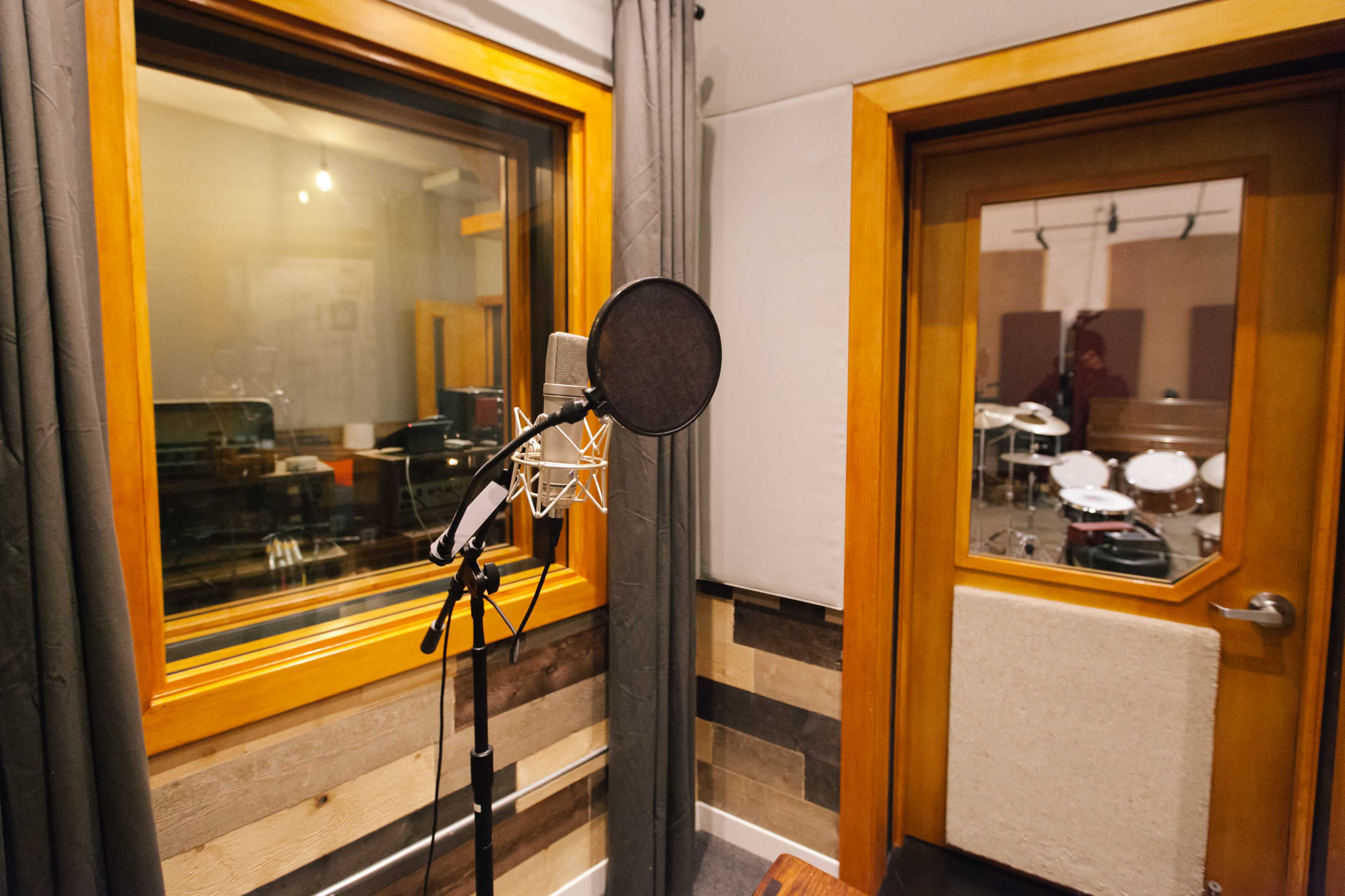 A recording studio with a microphone on a stand in the foreground and soundproof glass windows separating it from an area with musical instruments visible in the background.