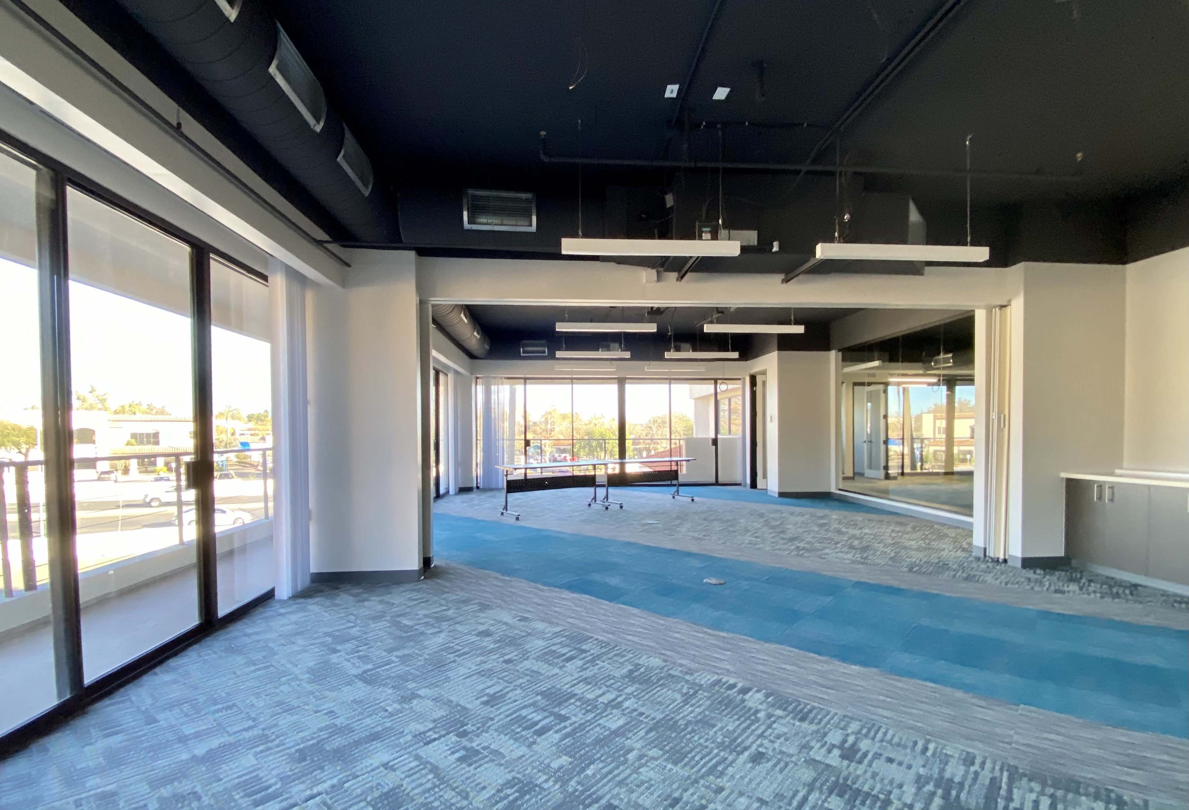 An empty modern office space features large windows, a conference table, and carpeted flooring in varying shades of blue and gray.
