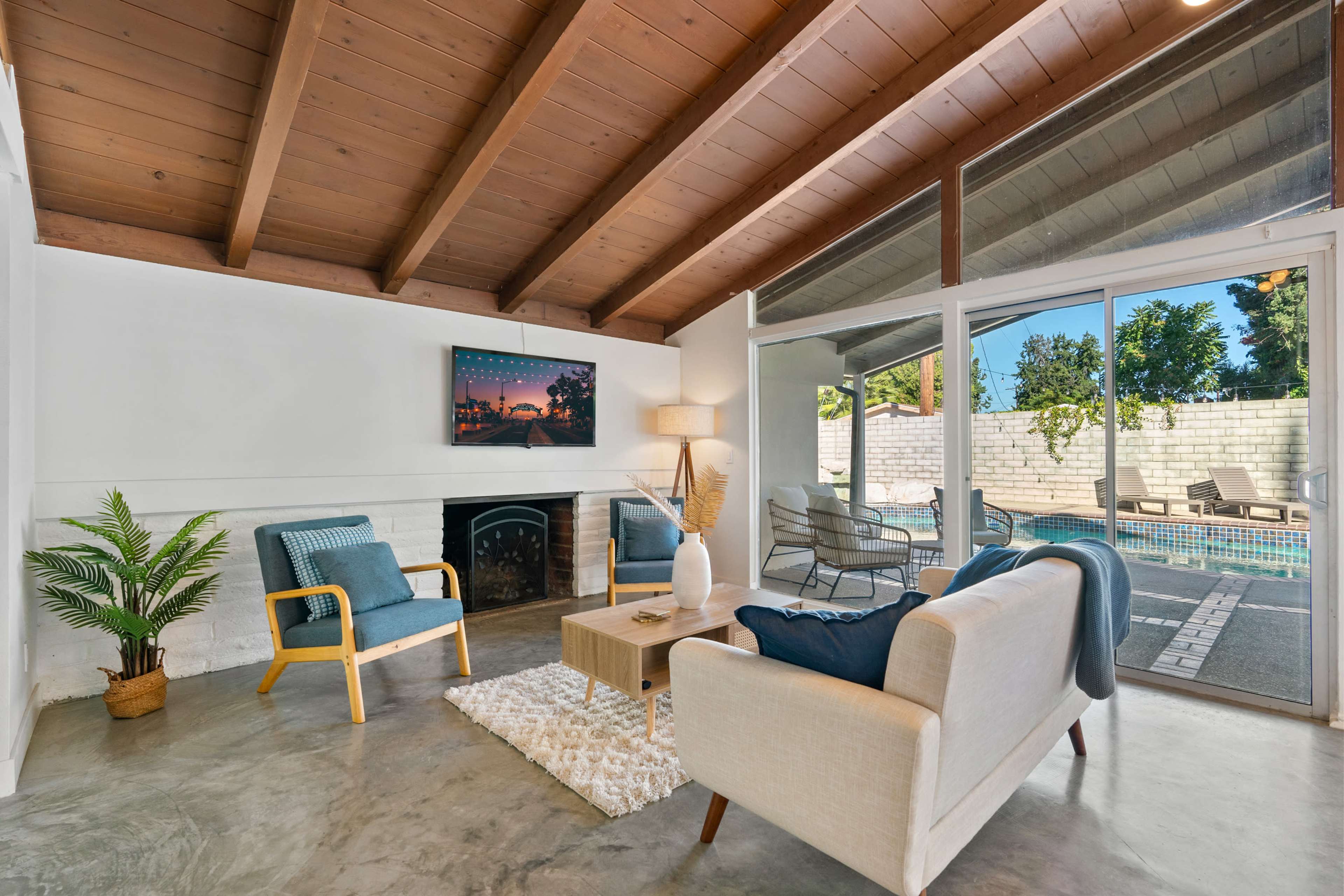 A modern living room with a fireplace, a television mounted on the wall, and large windows overlooking a pool area.