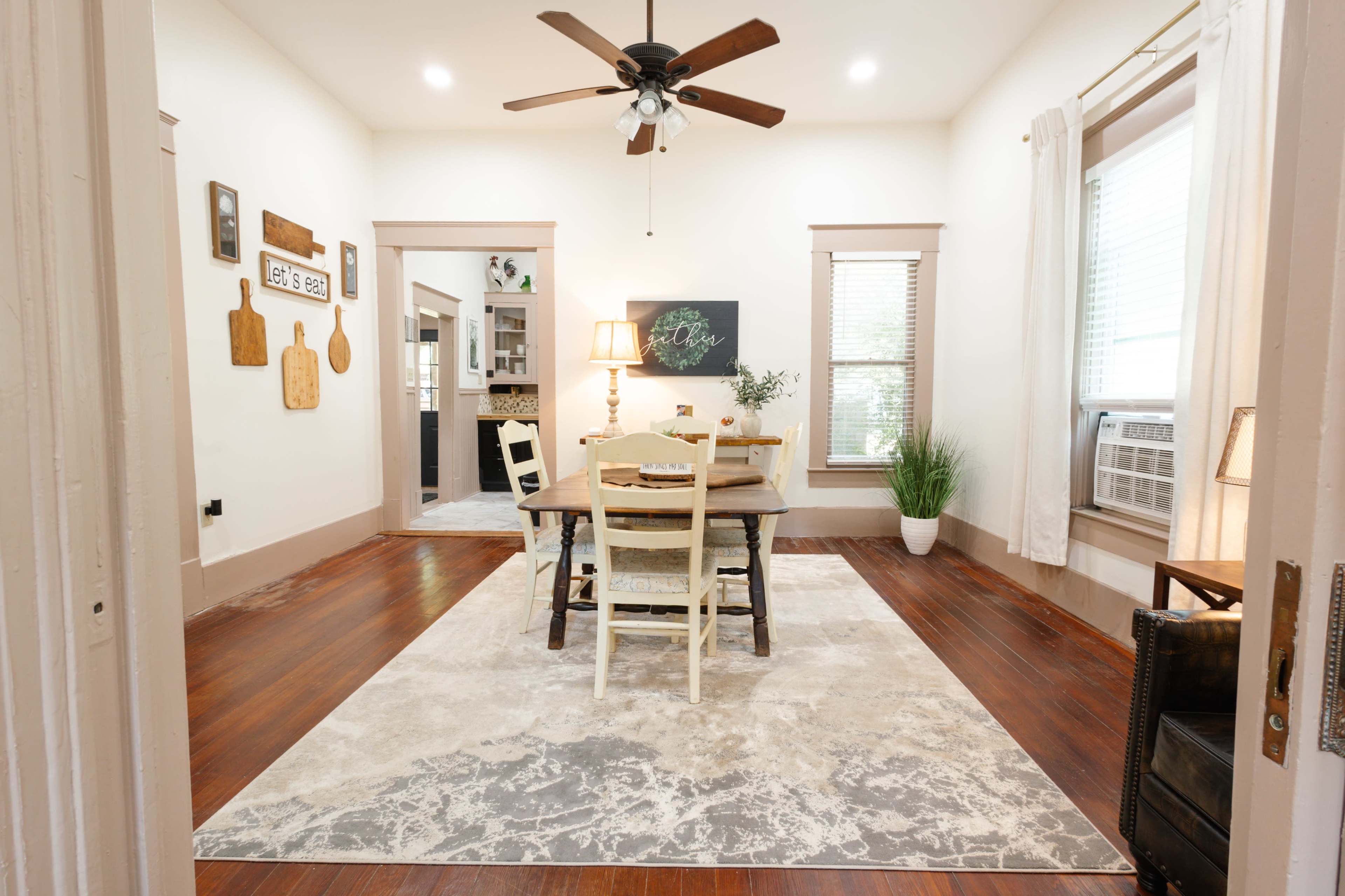 The image shows a dining area with a table and chairs on a large area rug, illuminated by natural light from the windows.