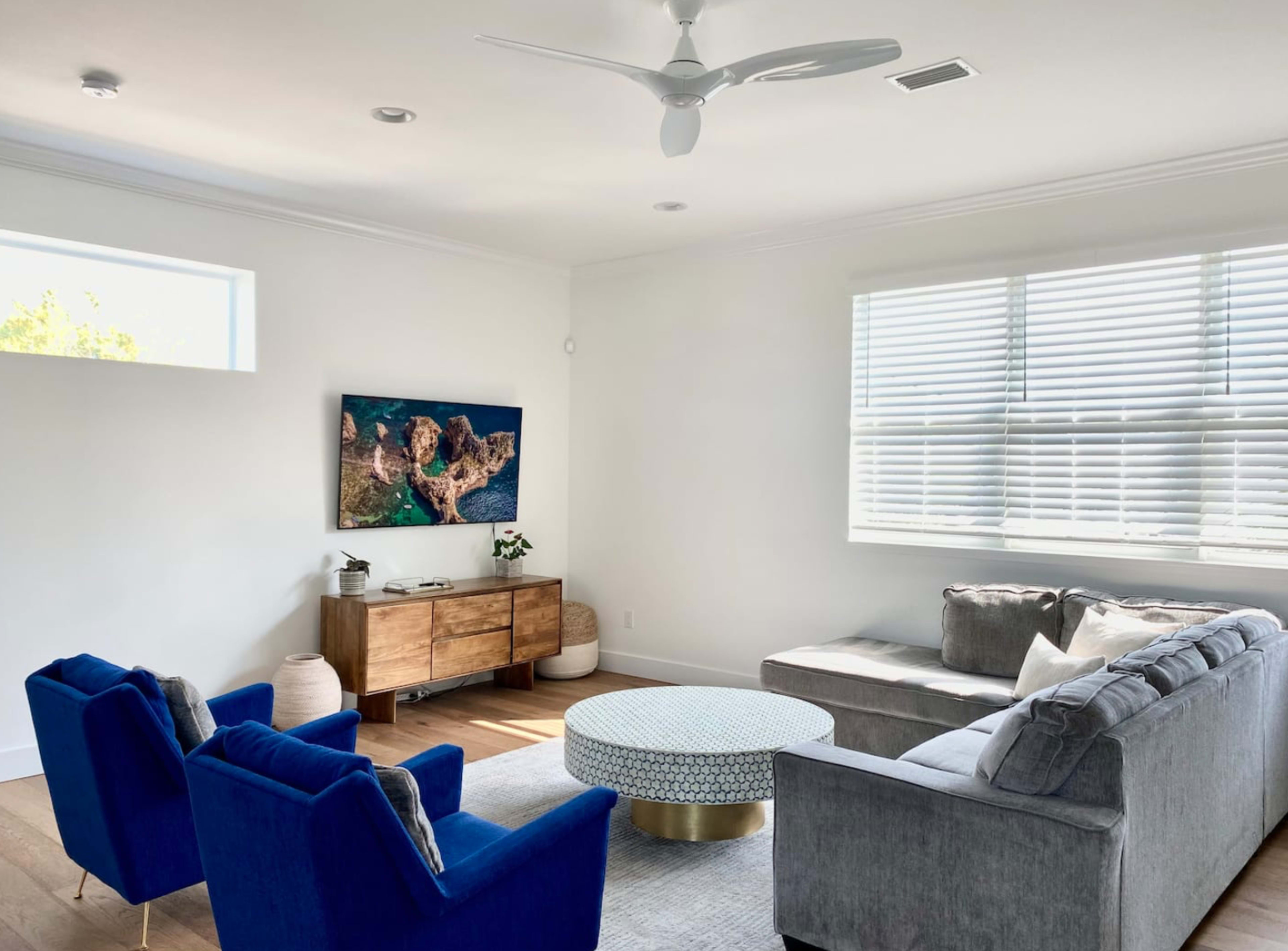 A modern living room with a sectional sofa, two blue chairs, a round coffee table, and a wall-mounted TV above a wooden console.