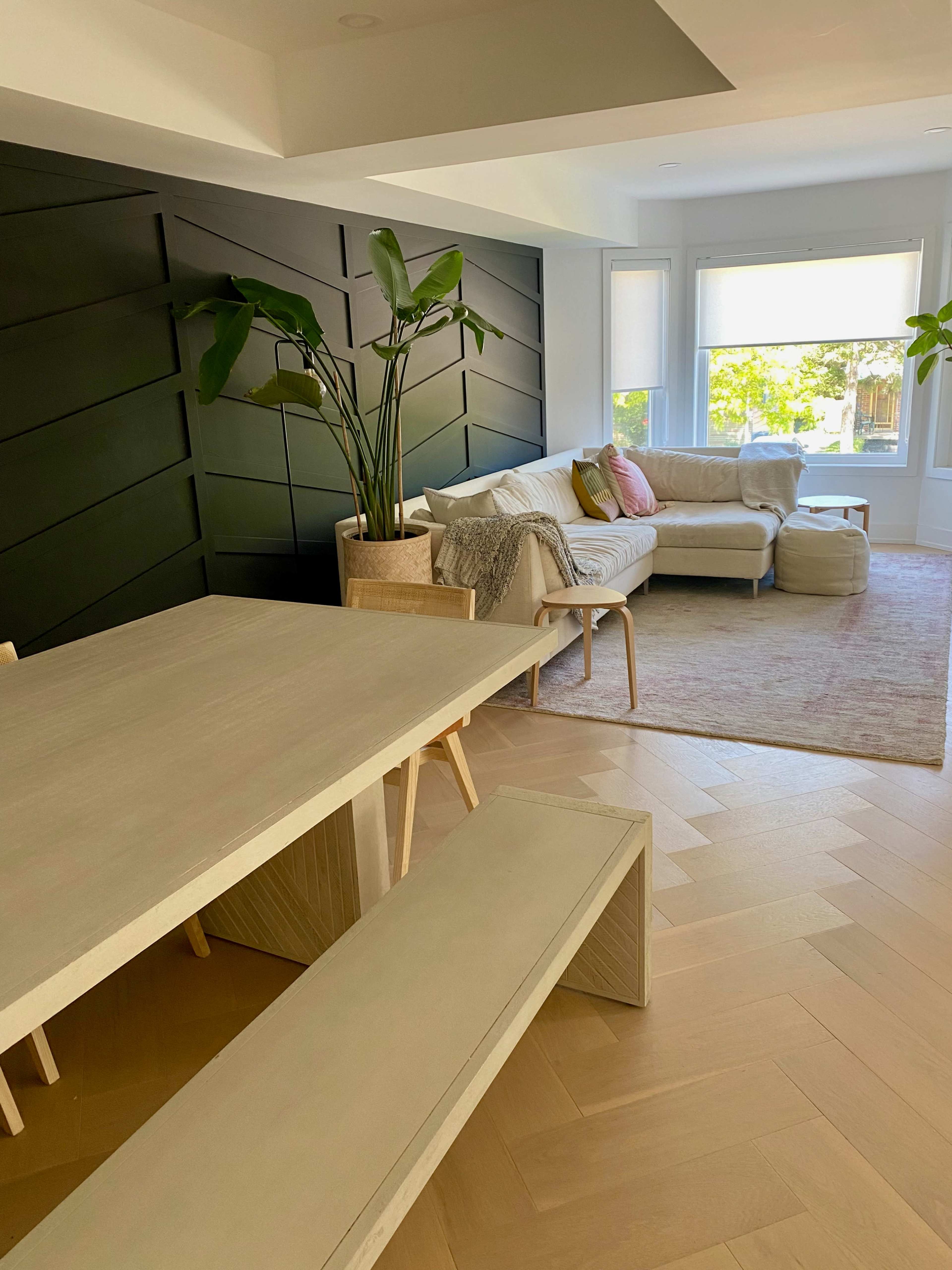 The image shows a modern living room and dining area with a minimalist table and bench, a textured green wall, and a light-colored sofa adorned with cushions.
