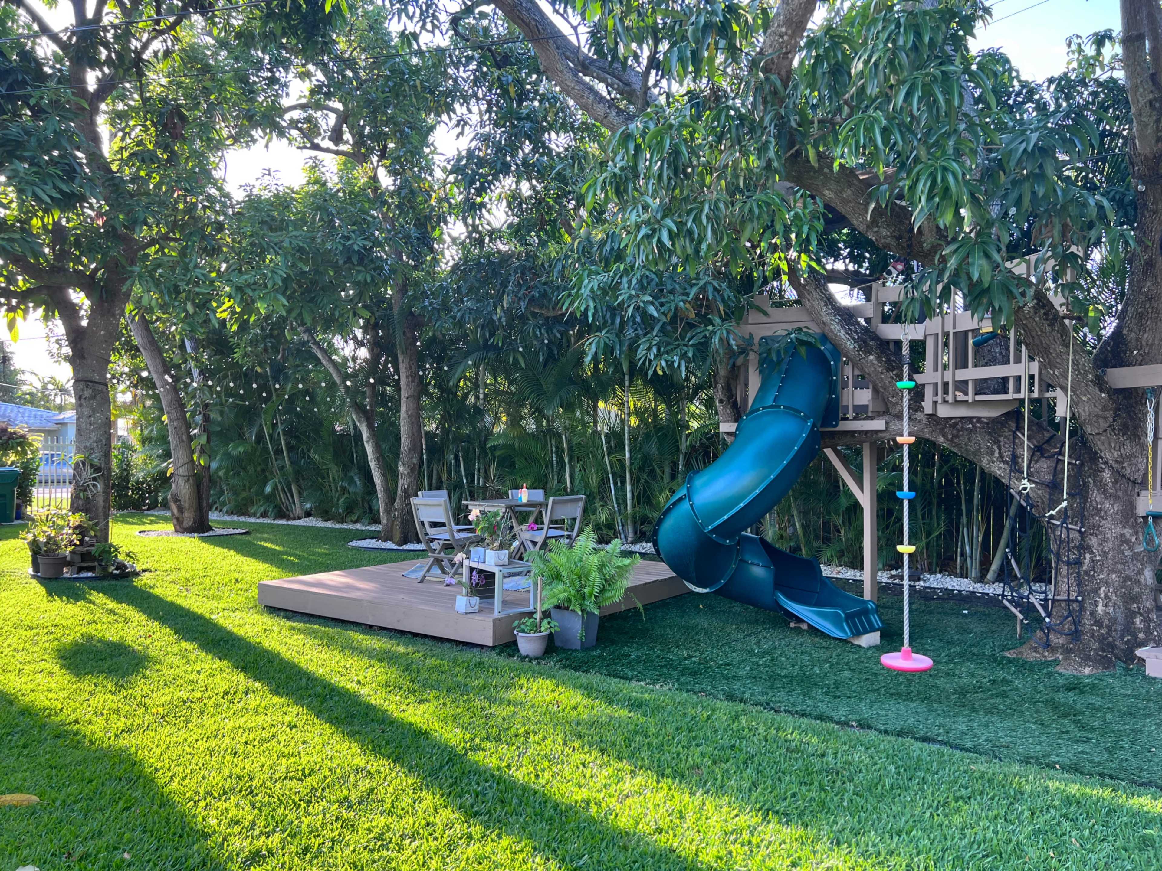 The image shows a backyard with a green slide attached to a treehouse, a wooden deck with seating, and well-manicured grass surrounded by trees.