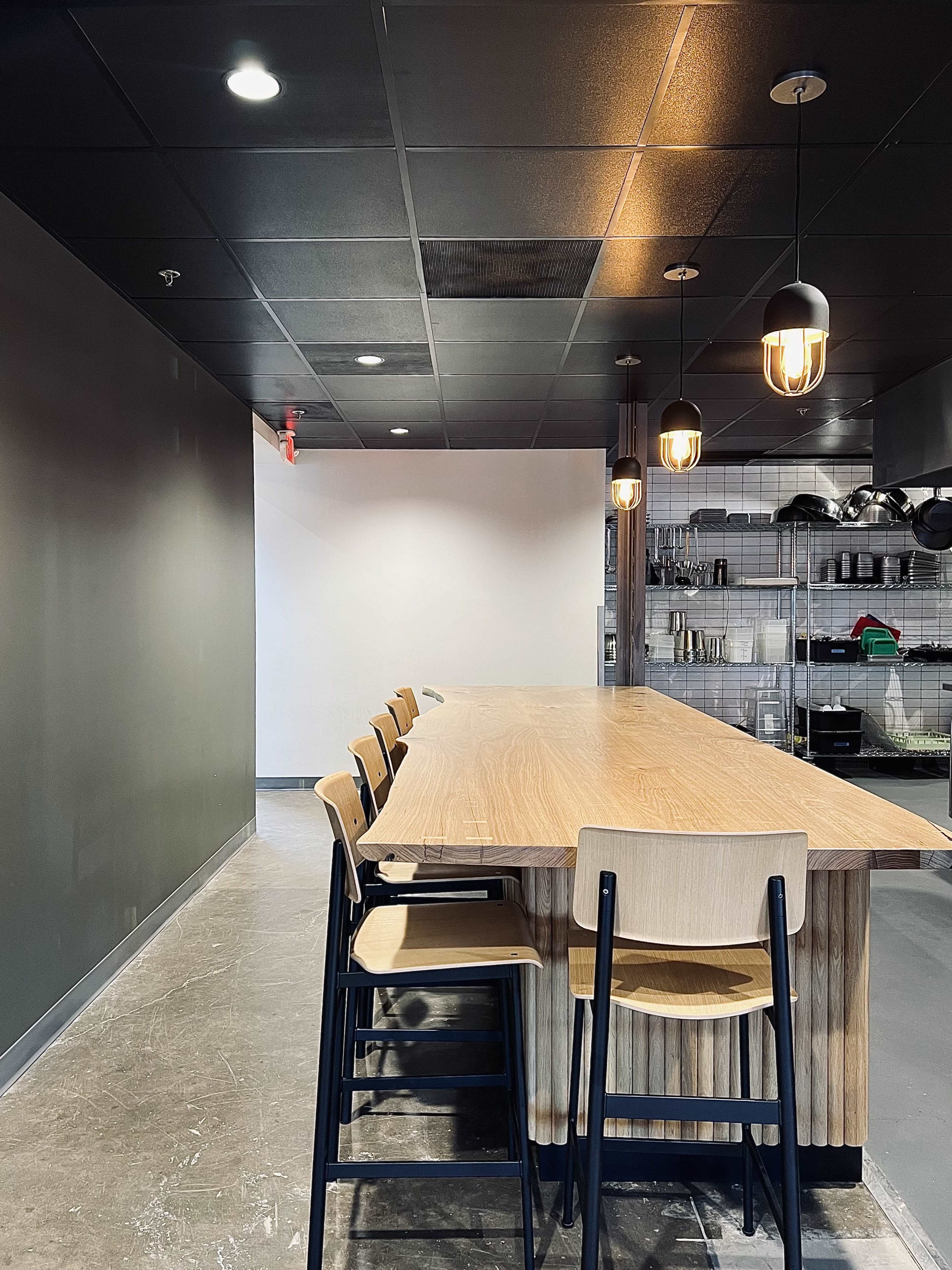 A long wooden table with black chairs is set against a wall in a modern industrial-style kitchen space featuring pendant lighting and open shelving with kitchen supplies.