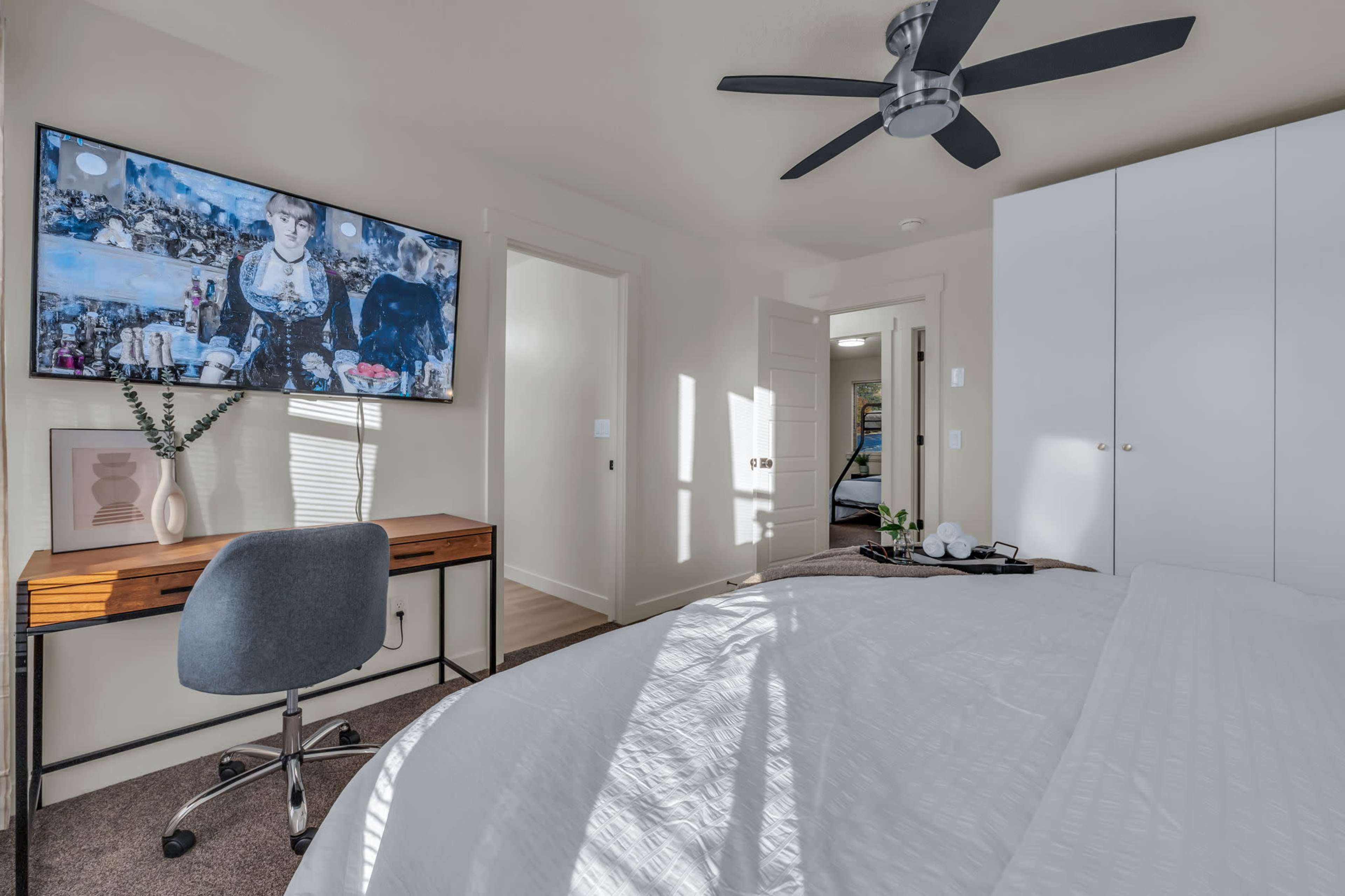 A modern bedroom with a large wall-mounted TV, a neatly made bed, a desk with a chair, and a closet in the background.