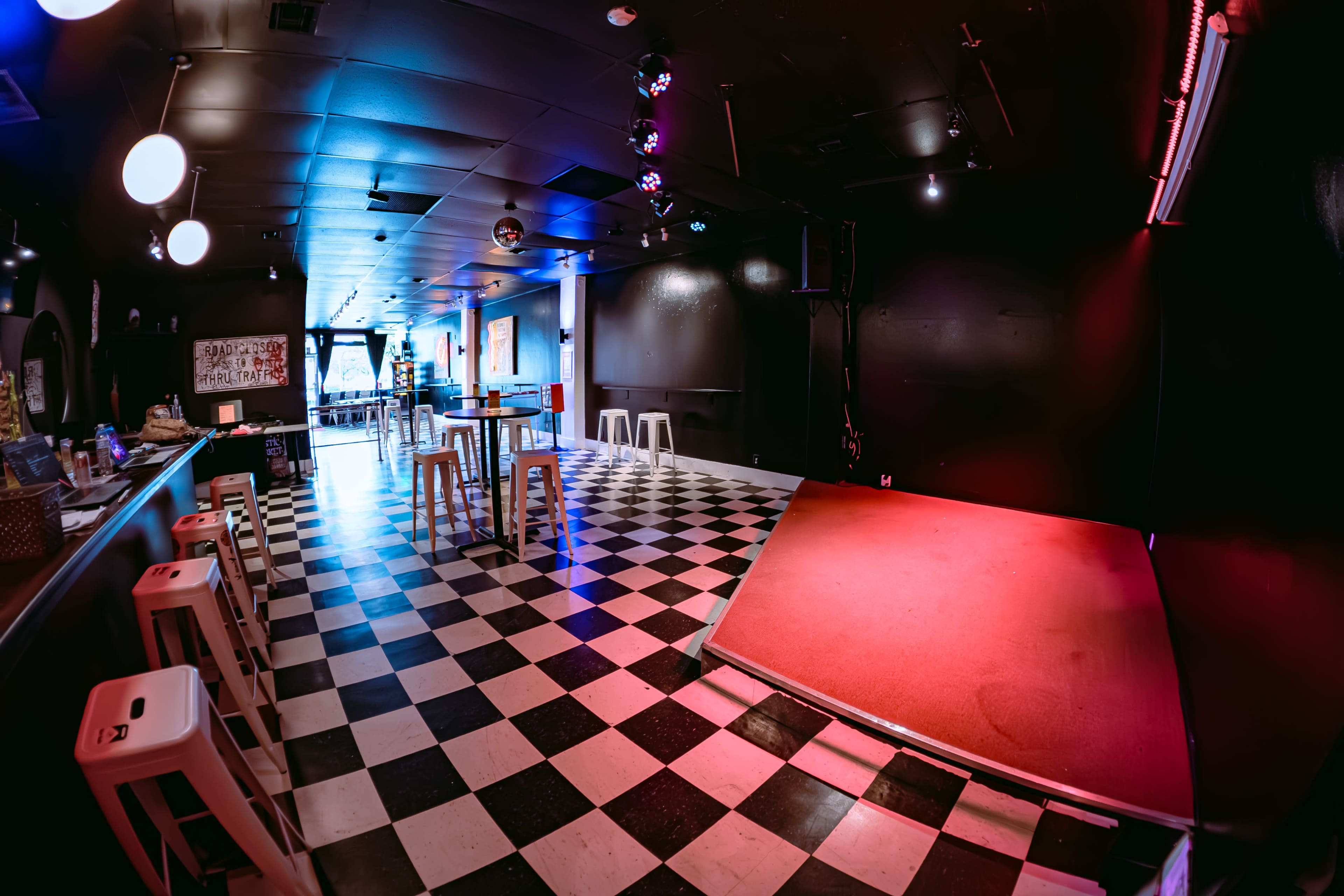 The image shows an empty, dimly lit venue with a checkered floor, a small raised stage, and seating areas in the background.