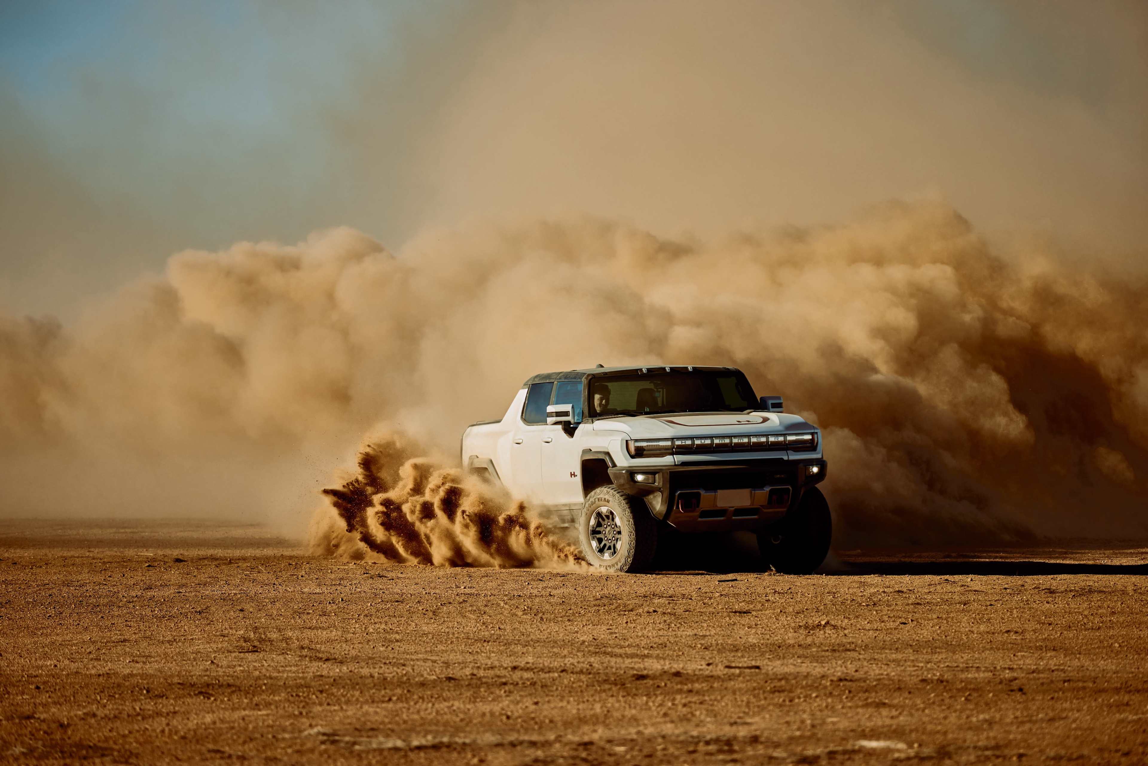 A white pickup truck kicks up a cloud of dust while driving on a dirt surface in a desert environment.