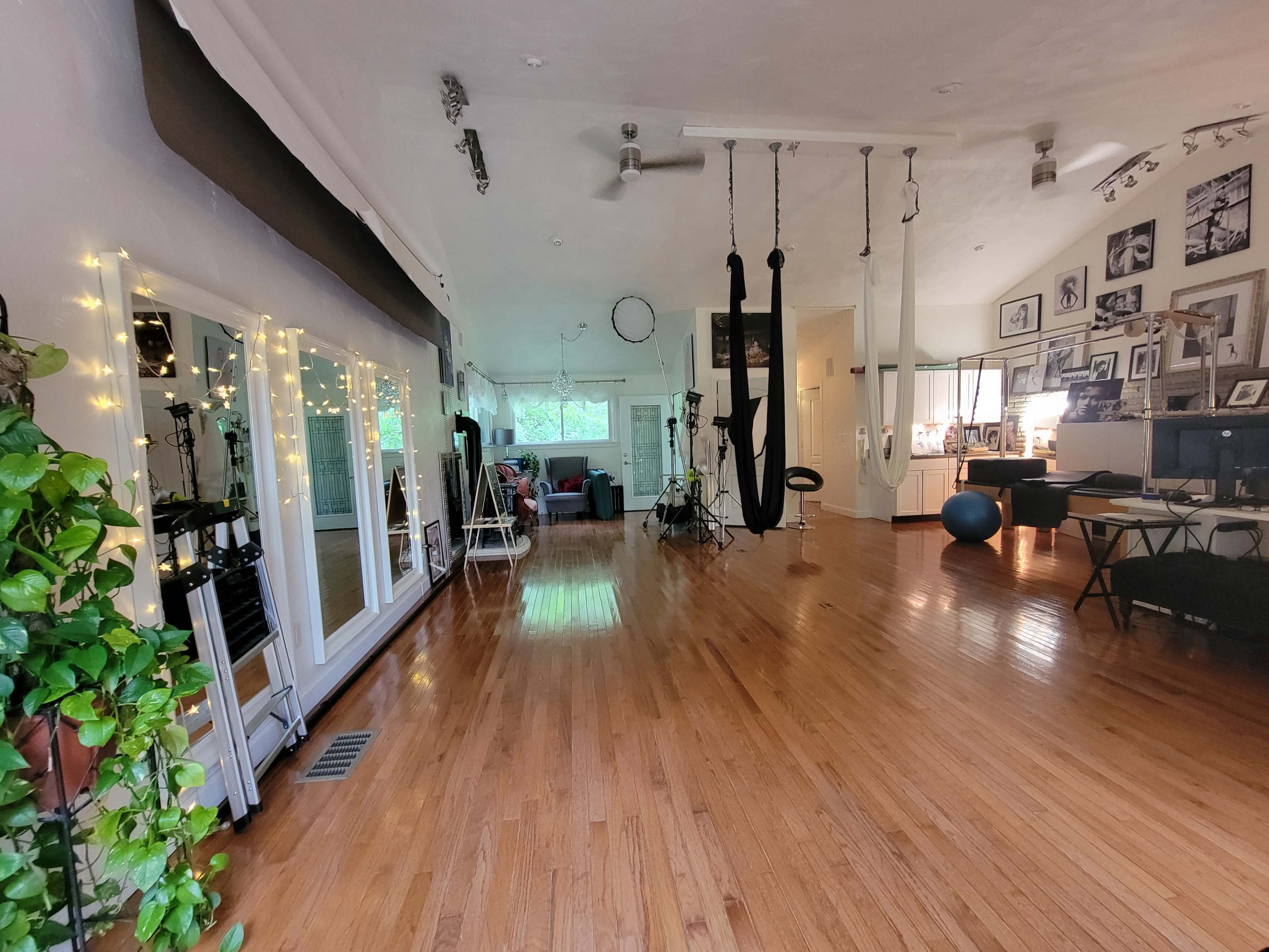 The image shows a spacious room with hardwood floors, large windows, various photographs on the walls, and gym equipment including aerial silks and exercise balls.