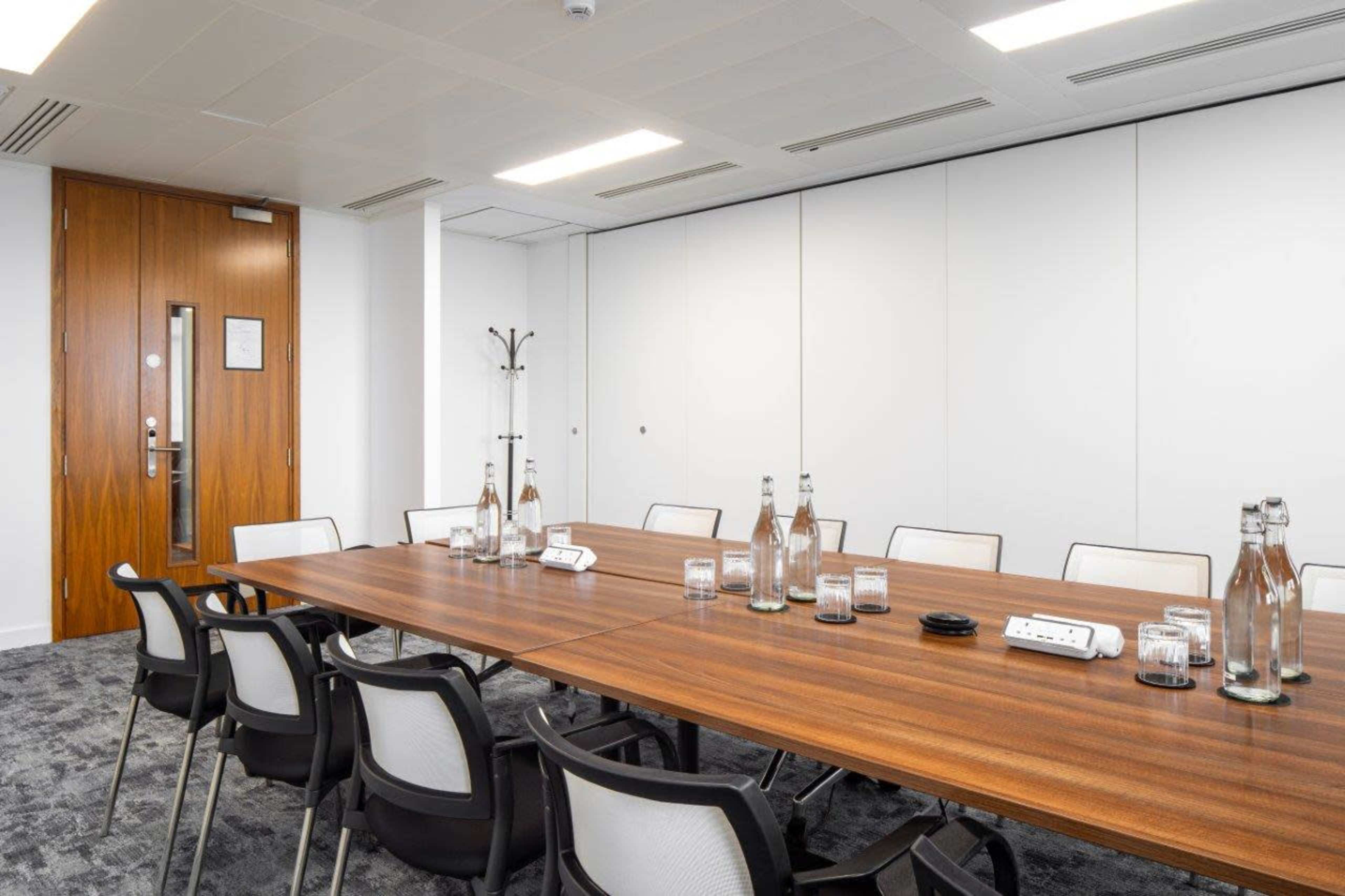 A large meeting room features a long wooden table surrounded by chairs, with water bottles and glasses placed on the table.