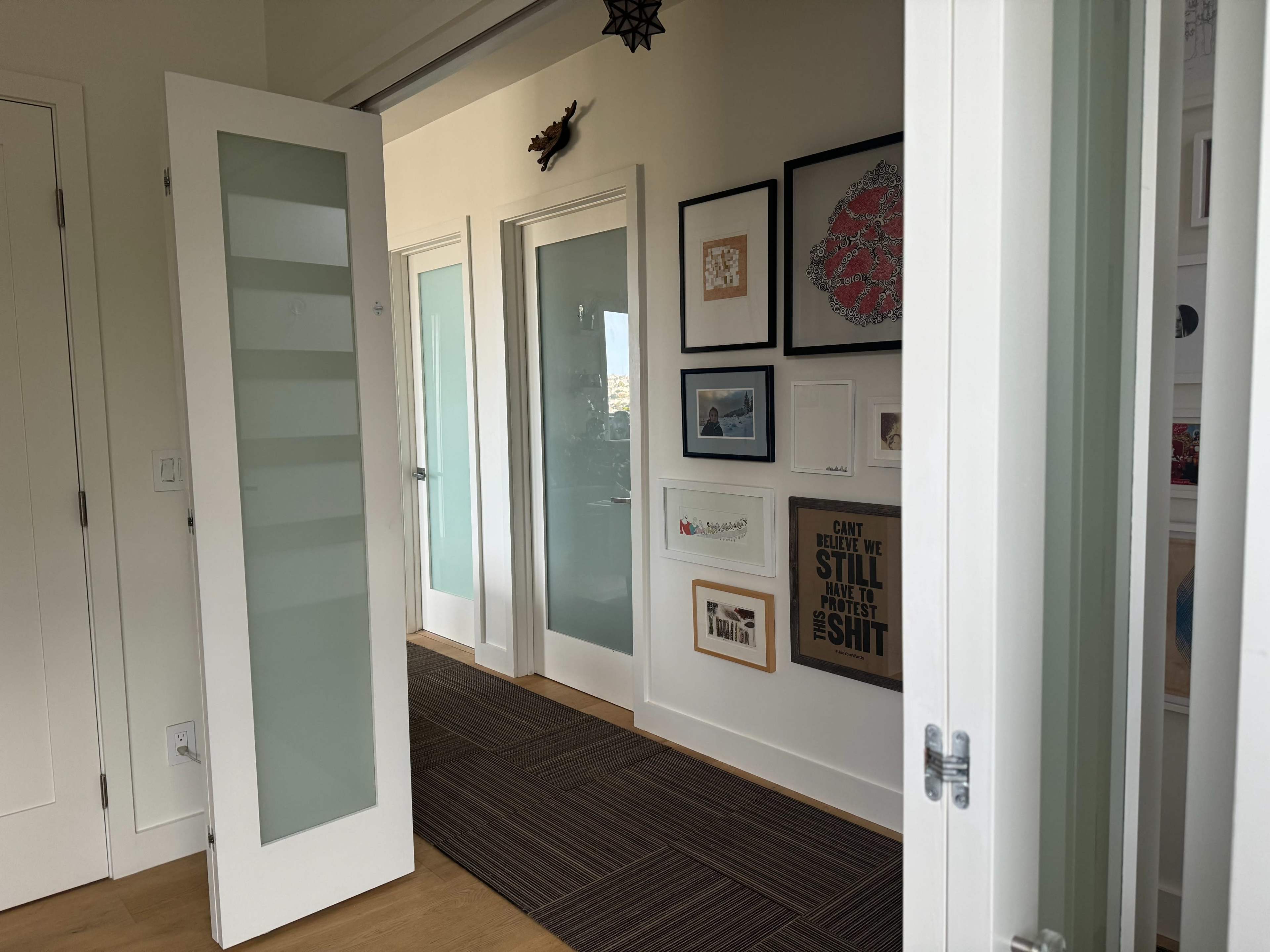 A modern hallway with glass-paneled doors, framed artworks on the walls, and a carpeted floor.
