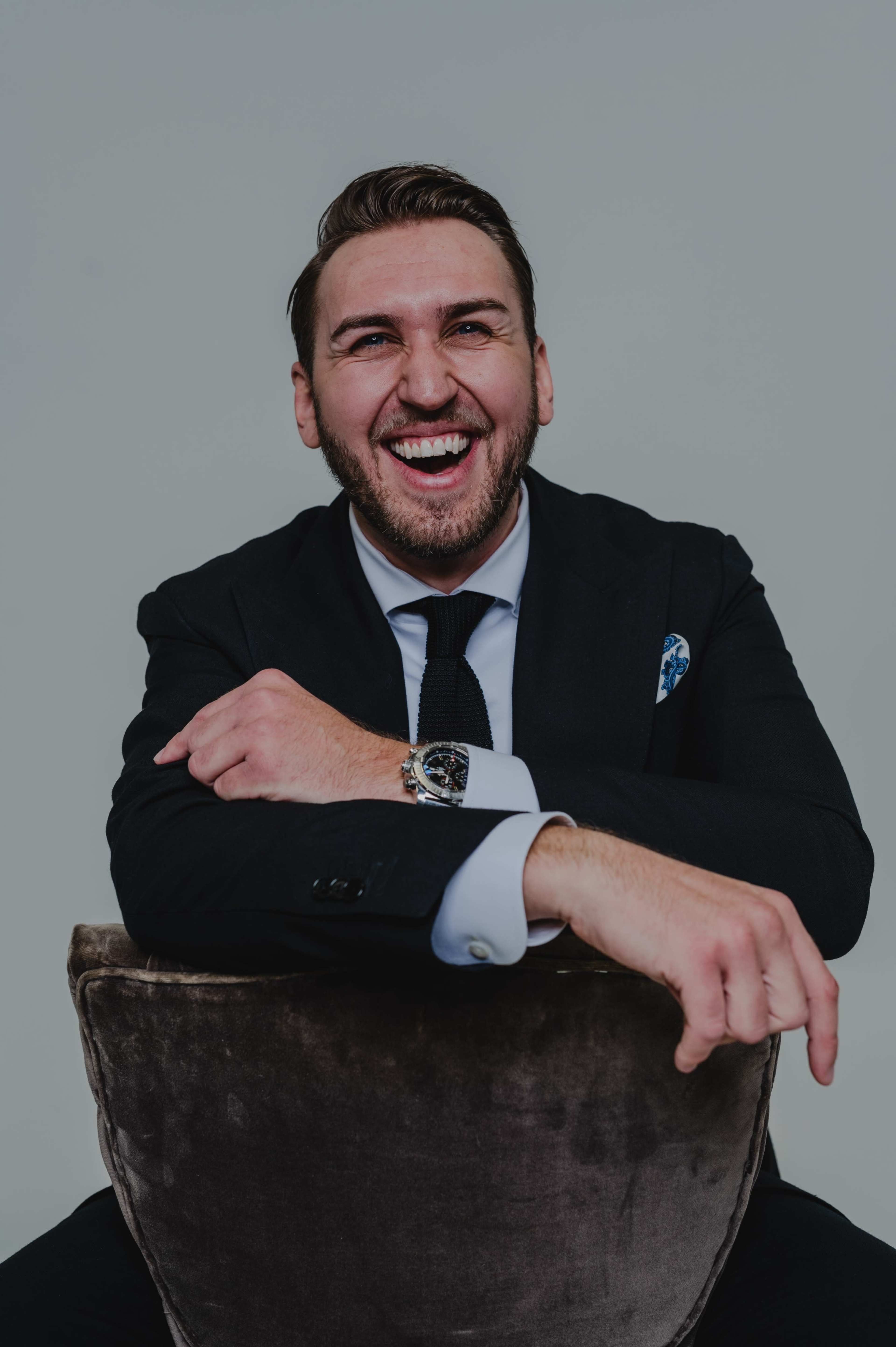 A man in a black suit laughs while sitting on a velvet chair.