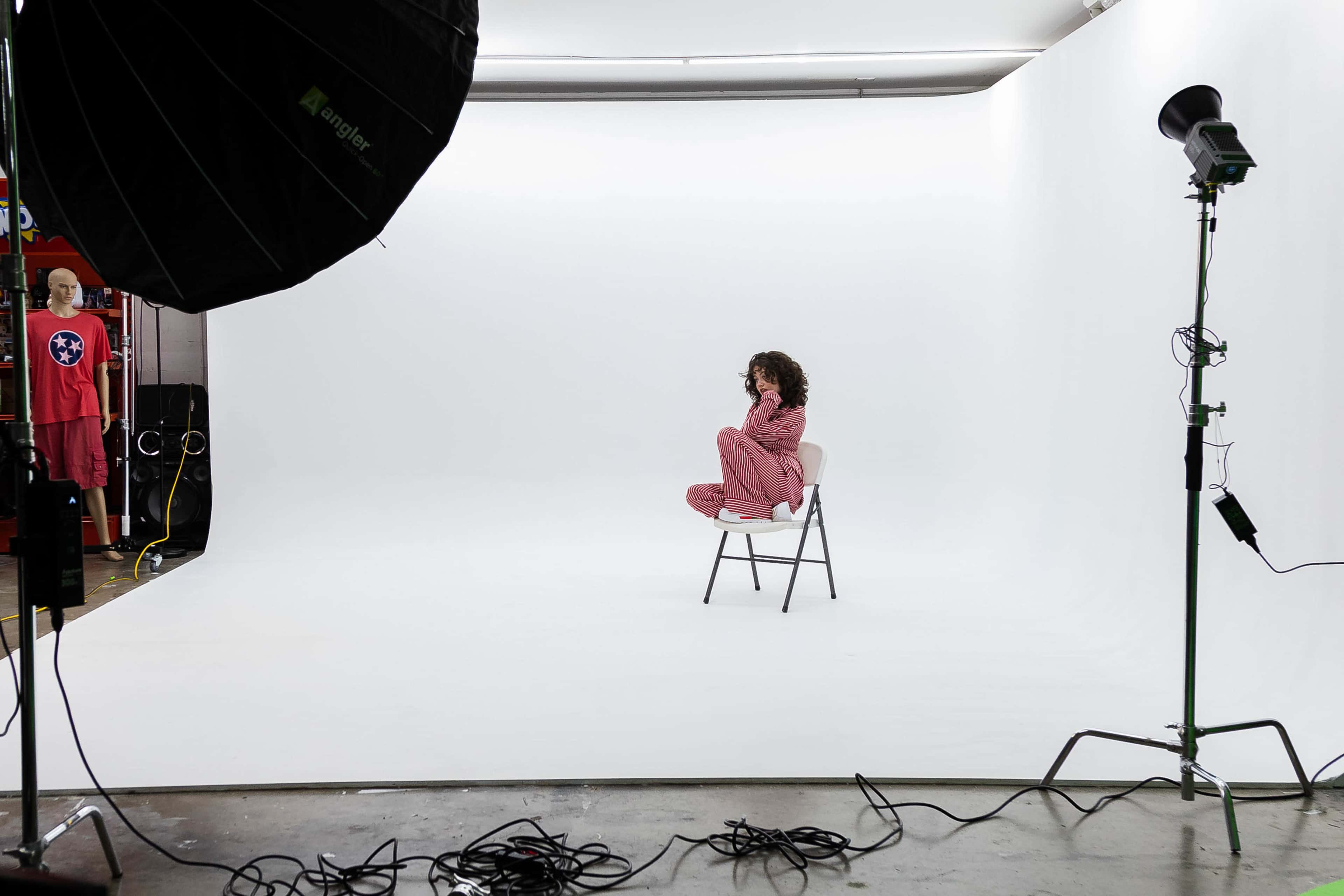 A person sits on a chair in a minimalist studio setting with a white backdrop, while a figure in a red outfit stands in the background.