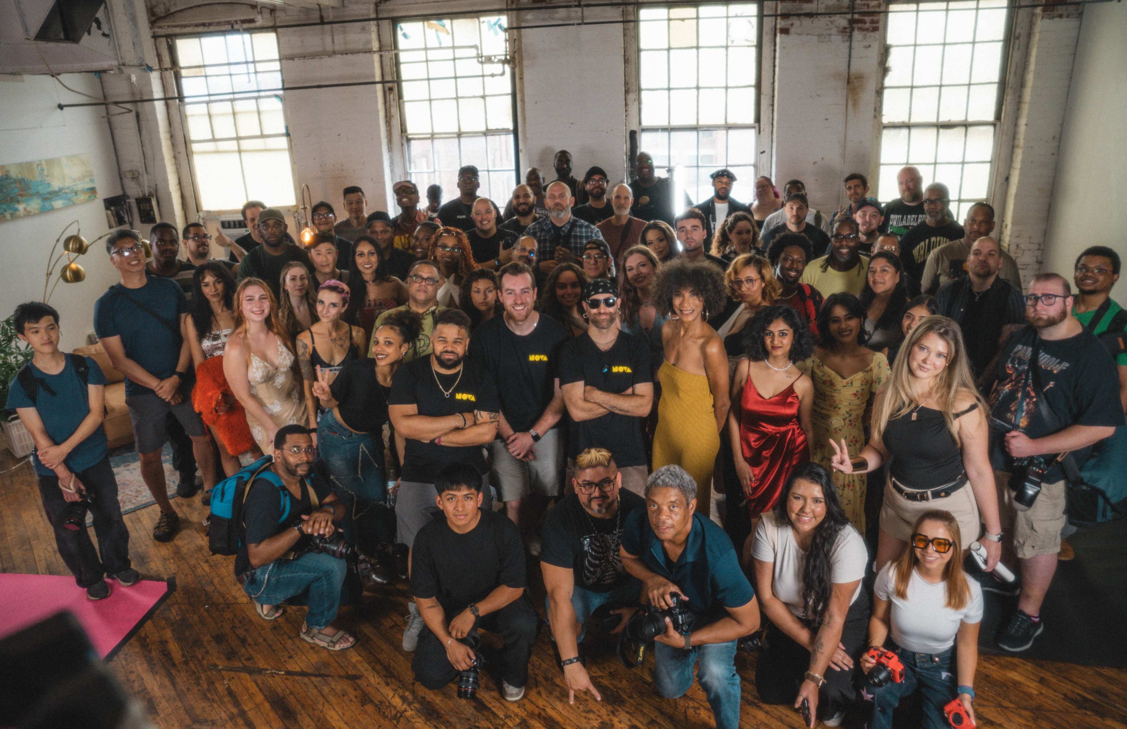 A large group of diverse people poses for a photo in an industrial-style studio with large windows.