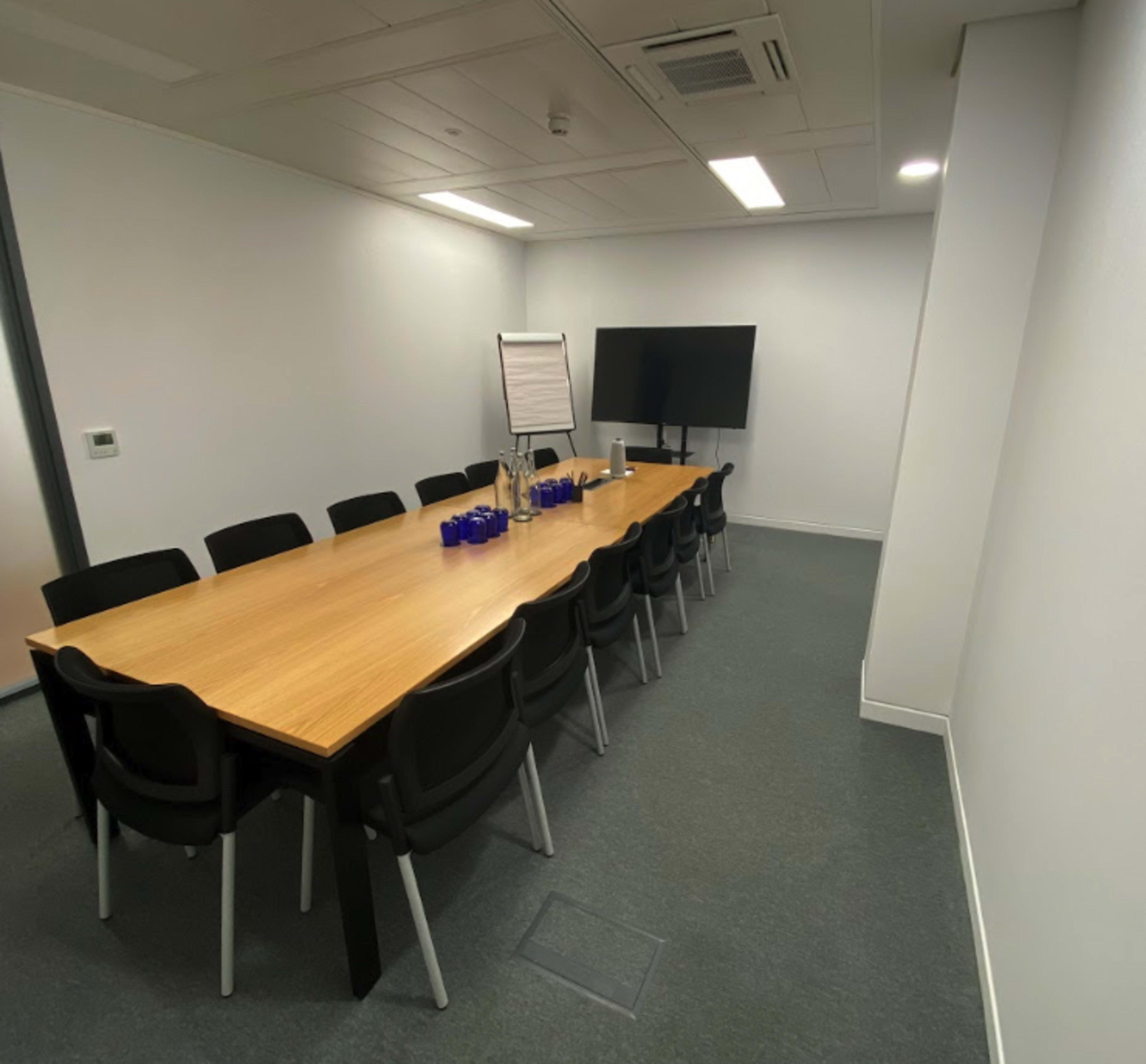 A conference room features a long wooden table surrounded by black chairs, with a whiteboard and a large screen on the walls.