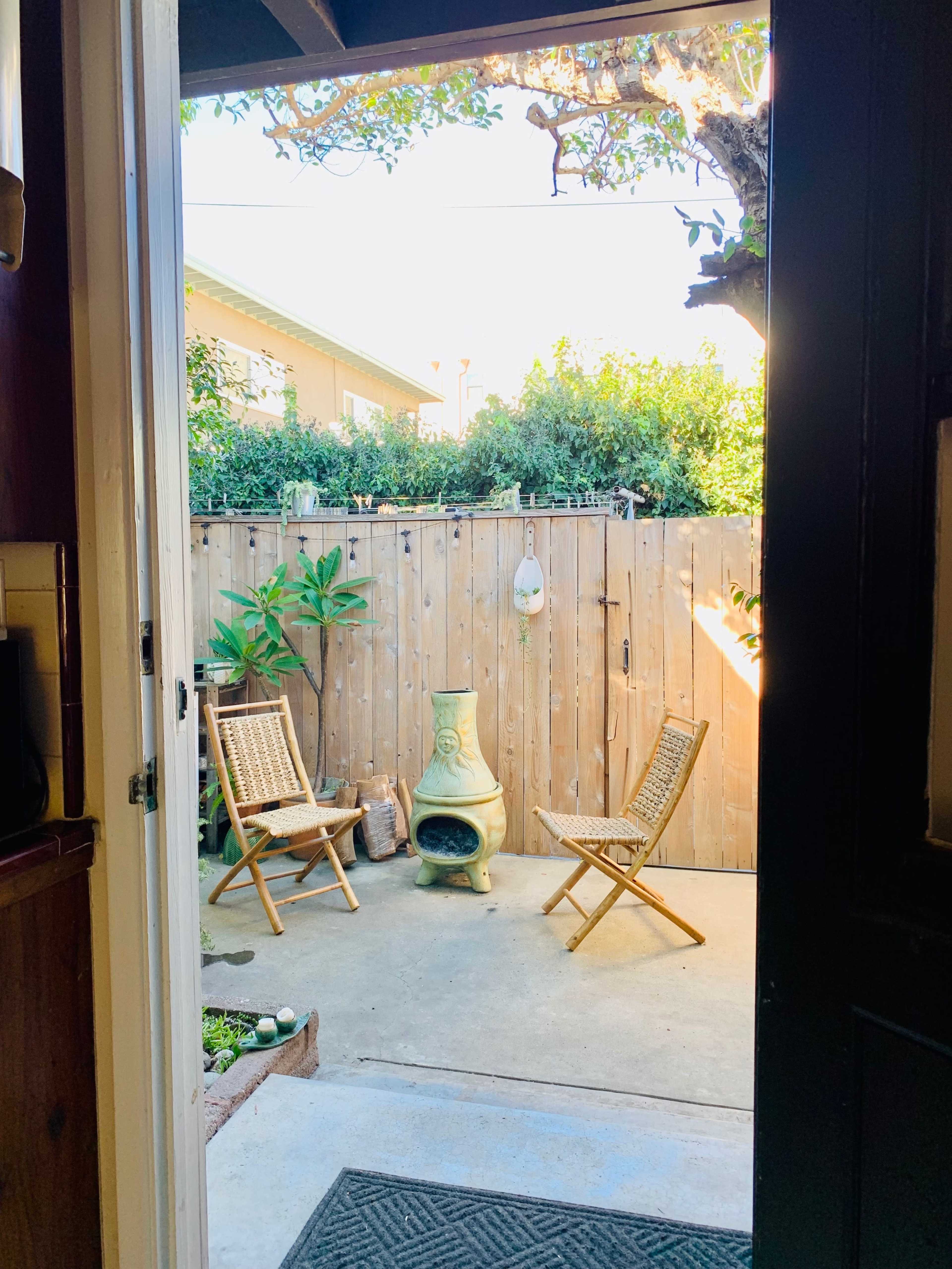 A view from an open door into a small outdoor patio with two wicker chairs and a green chiminea, surrounded by a wooden fence and plants.