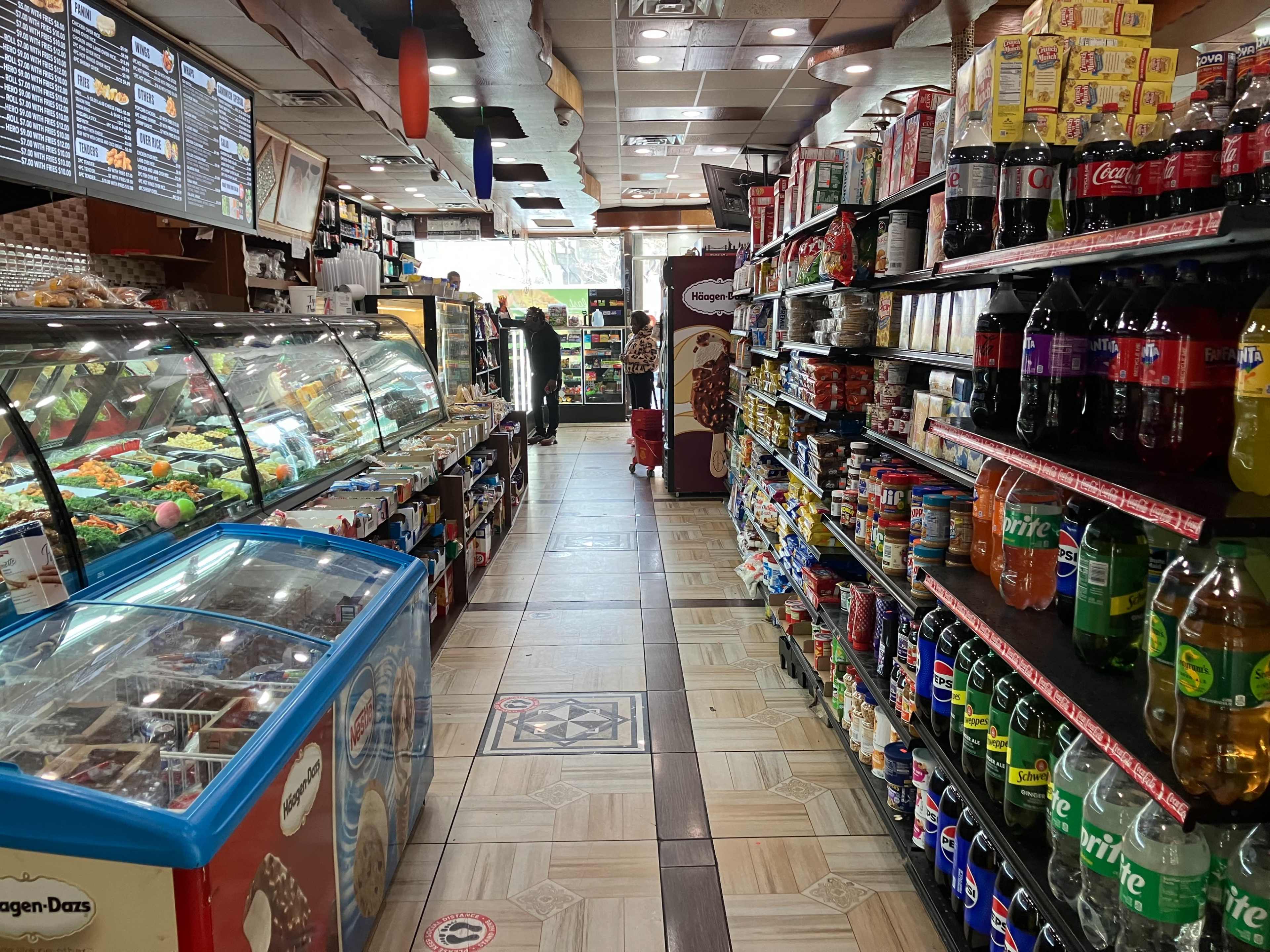 The interior of a convenience store with shelves stocked on both sides and a display case filled with food items at the front.