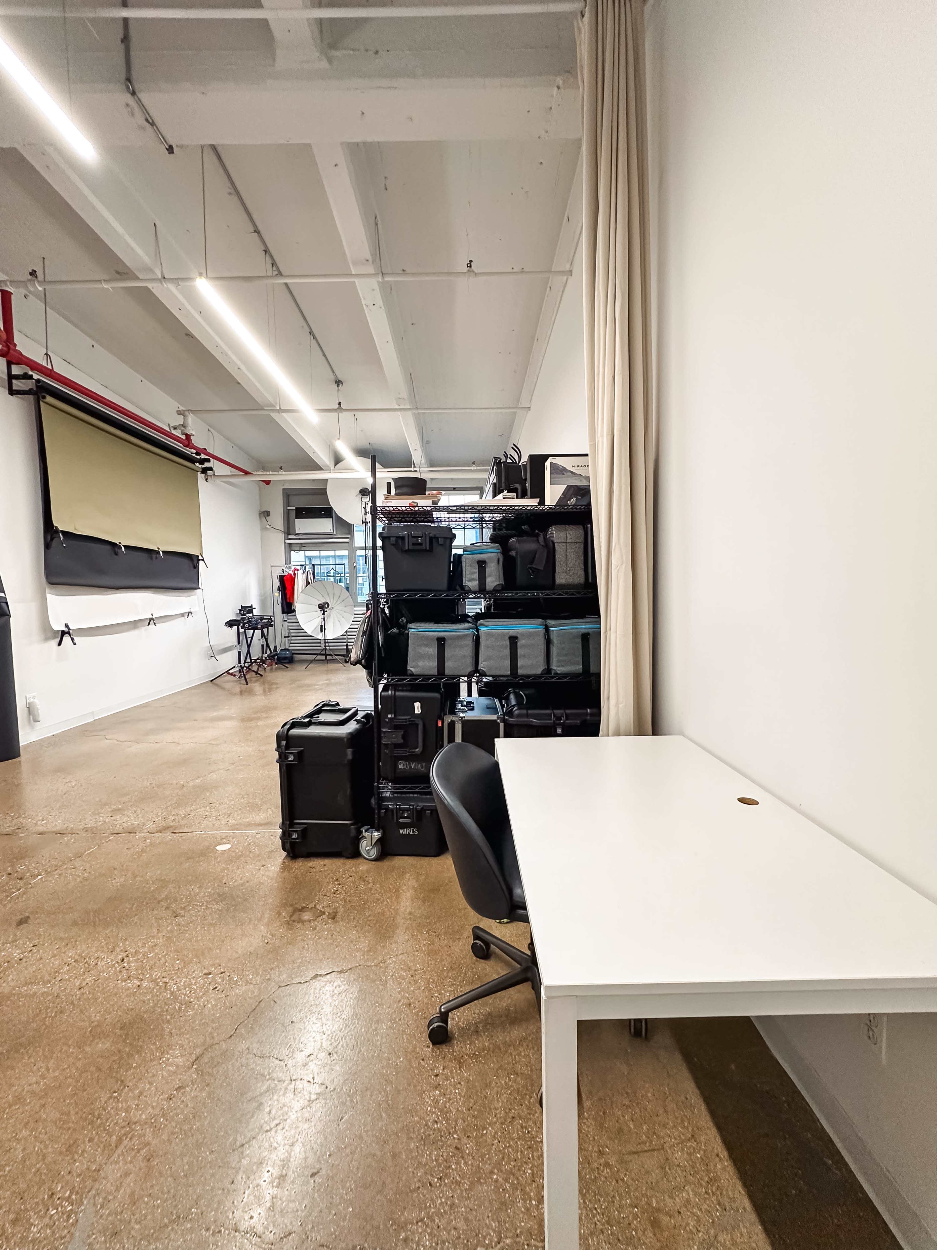 A spacious room with a white desk and a black chair next to a stack of storage cases, with a large screen and equipment in the background.
