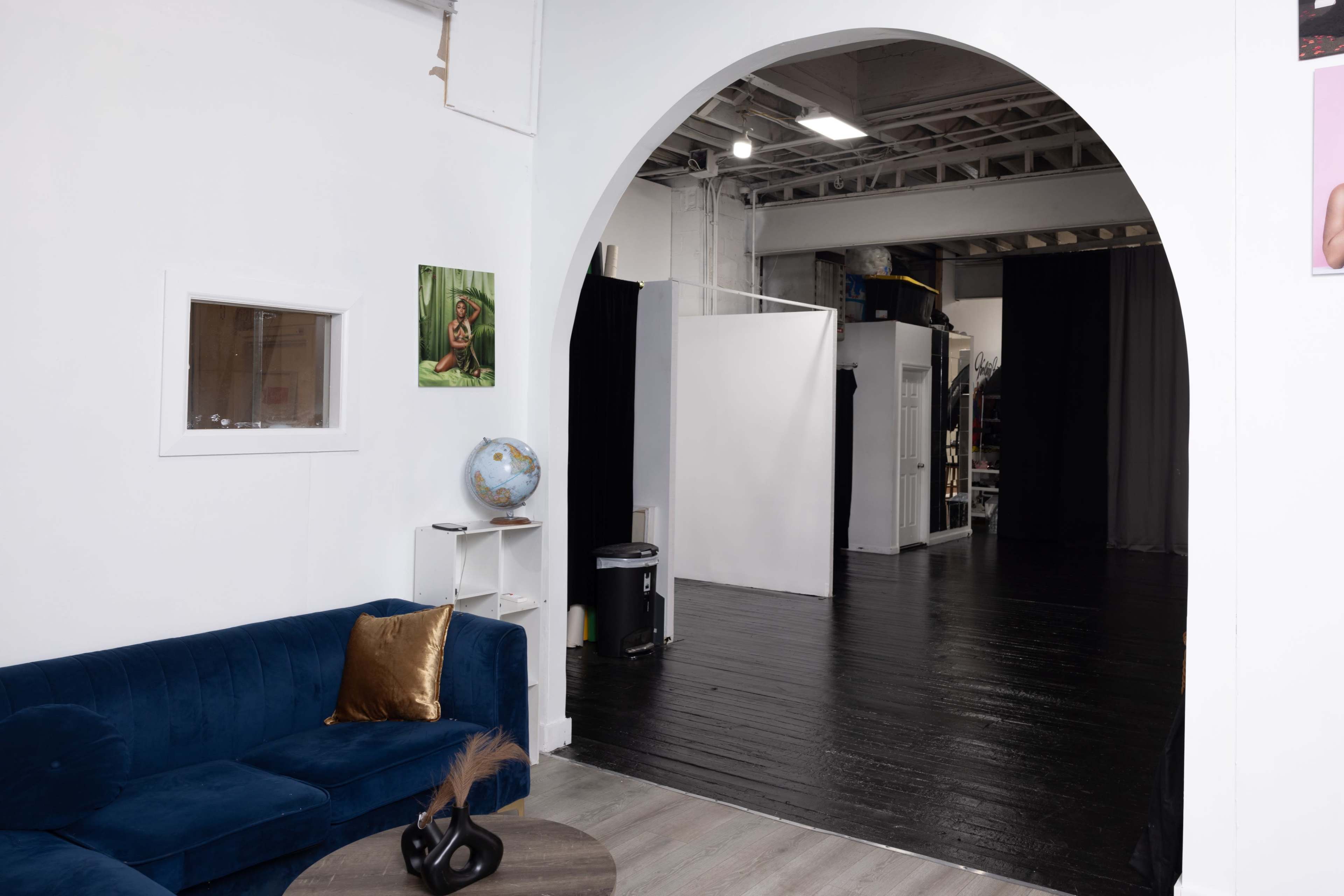 The image shows a modern studio space featuring a blue couch, a globe on a side table, and a large archway leading to a darkened area with a white backdrop.