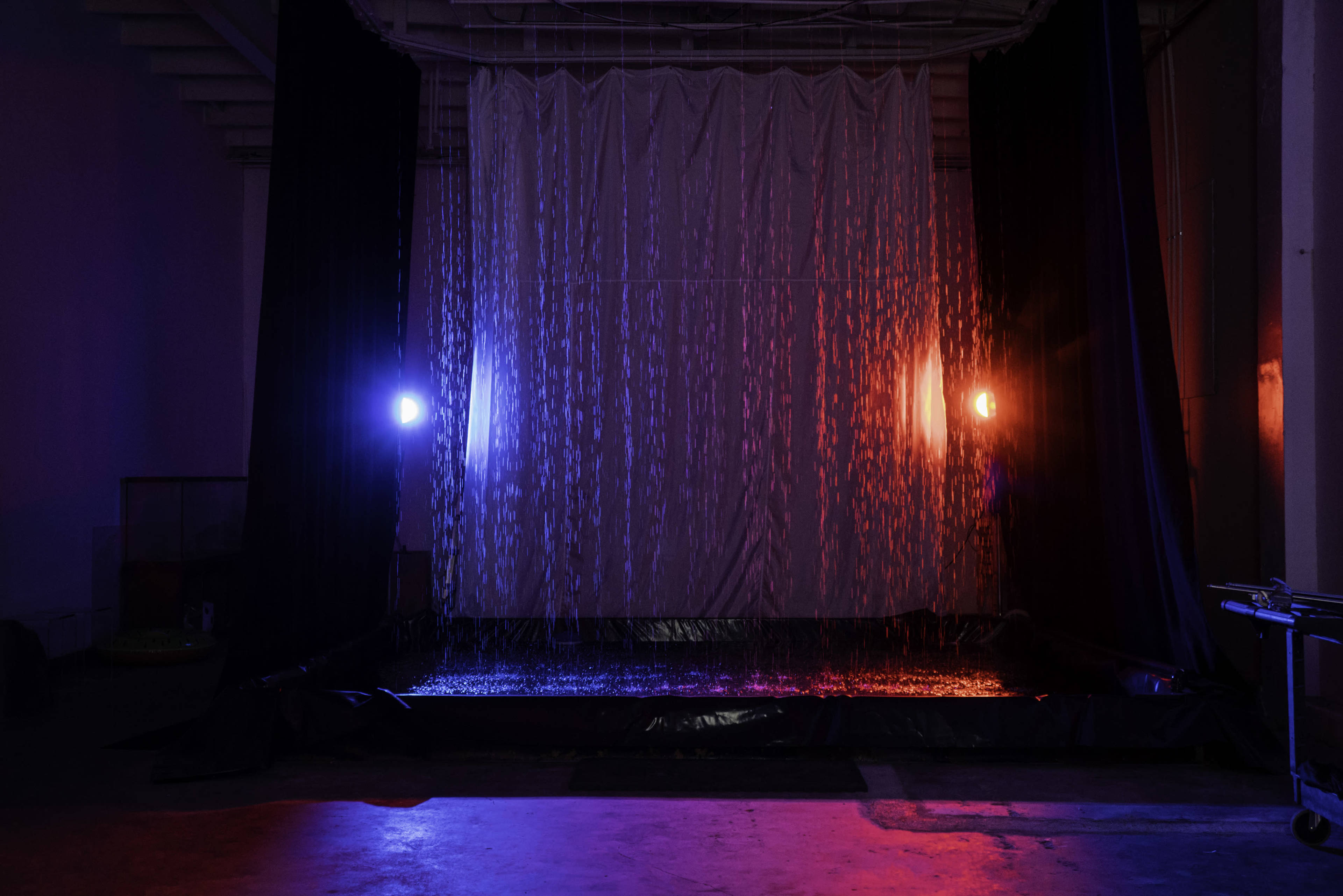 A curtain of water flows down from a suspended fabric backdrop, illuminated by red and blue lights in a darkened space.