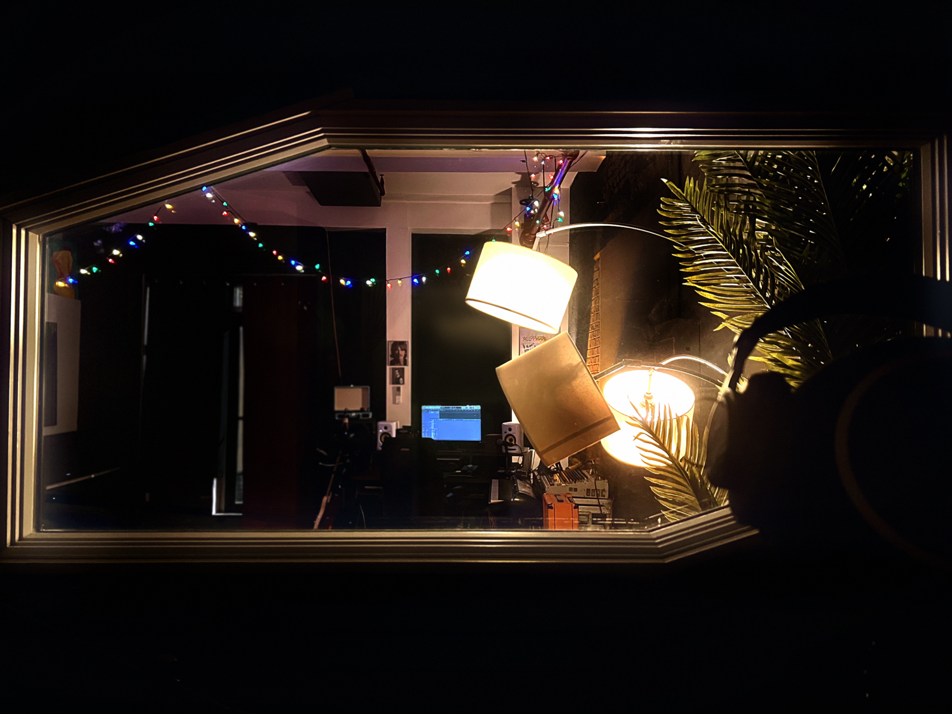 The image shows a studio room visible through a decorative window frame, featuring illuminated lamps, a computer setup, and colorful string lights in the background.