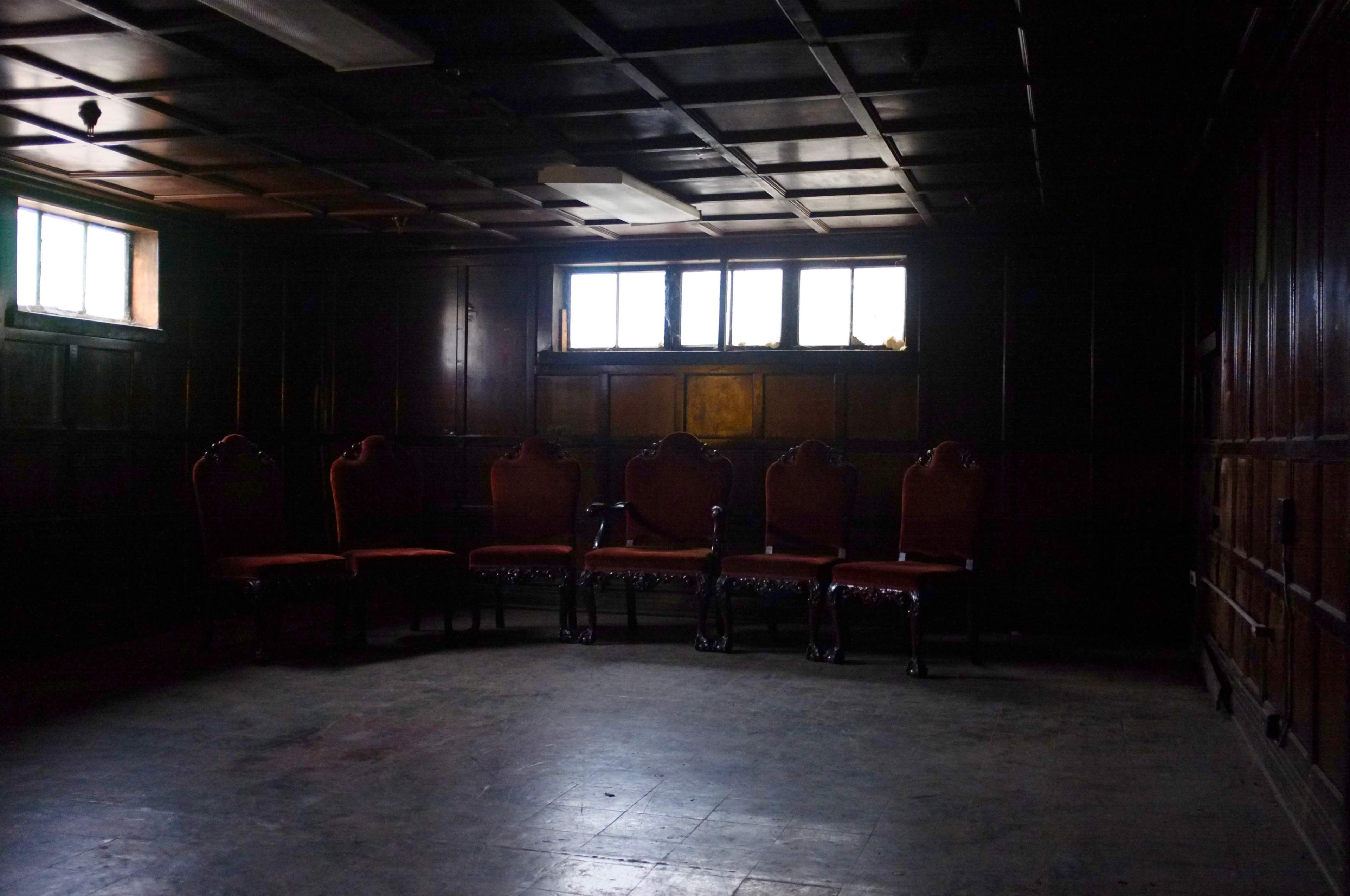 A dimly lit room features a row of five ornate chairs against a dark wooden wall, with two small windows allowing faint light to enter.