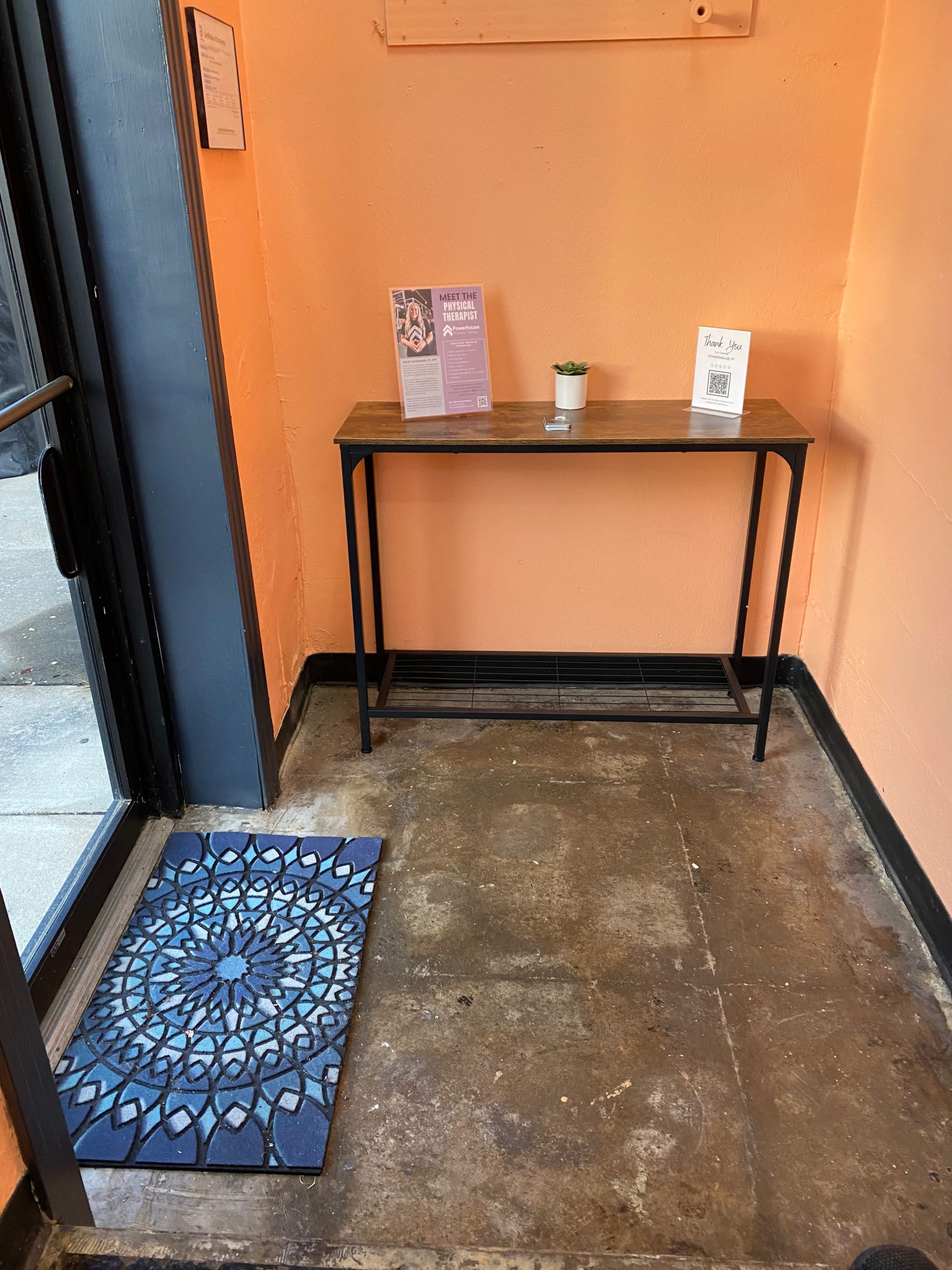 The image shows a small entryway with an orange wall, a wooden table against it displaying pamphlets, and a blue patterned mat in front of an open door.