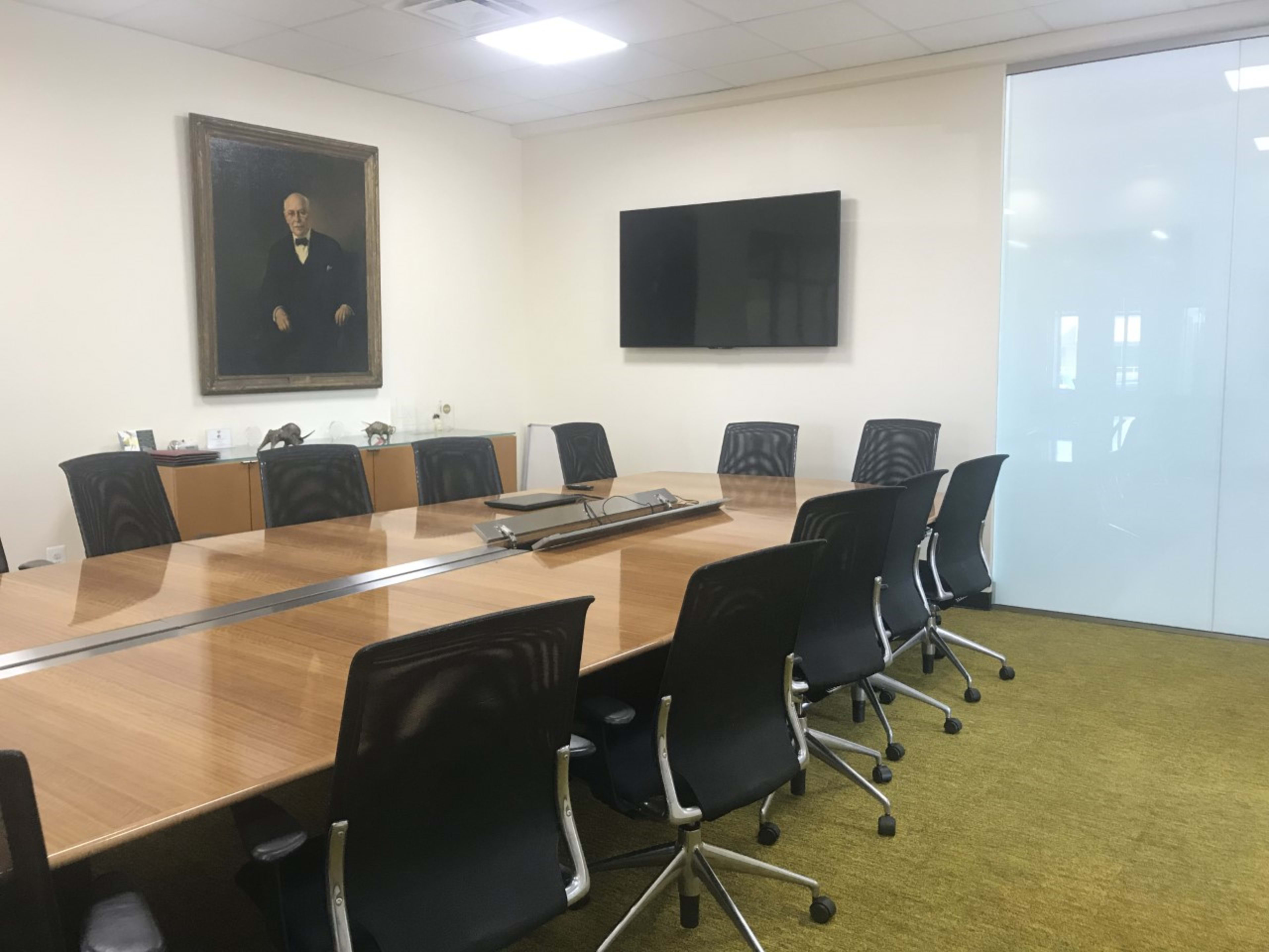 A modern conference room features a large wooden table surrounded by black chairs, a wall-mounted television, and a portrait hanging on the wall.
