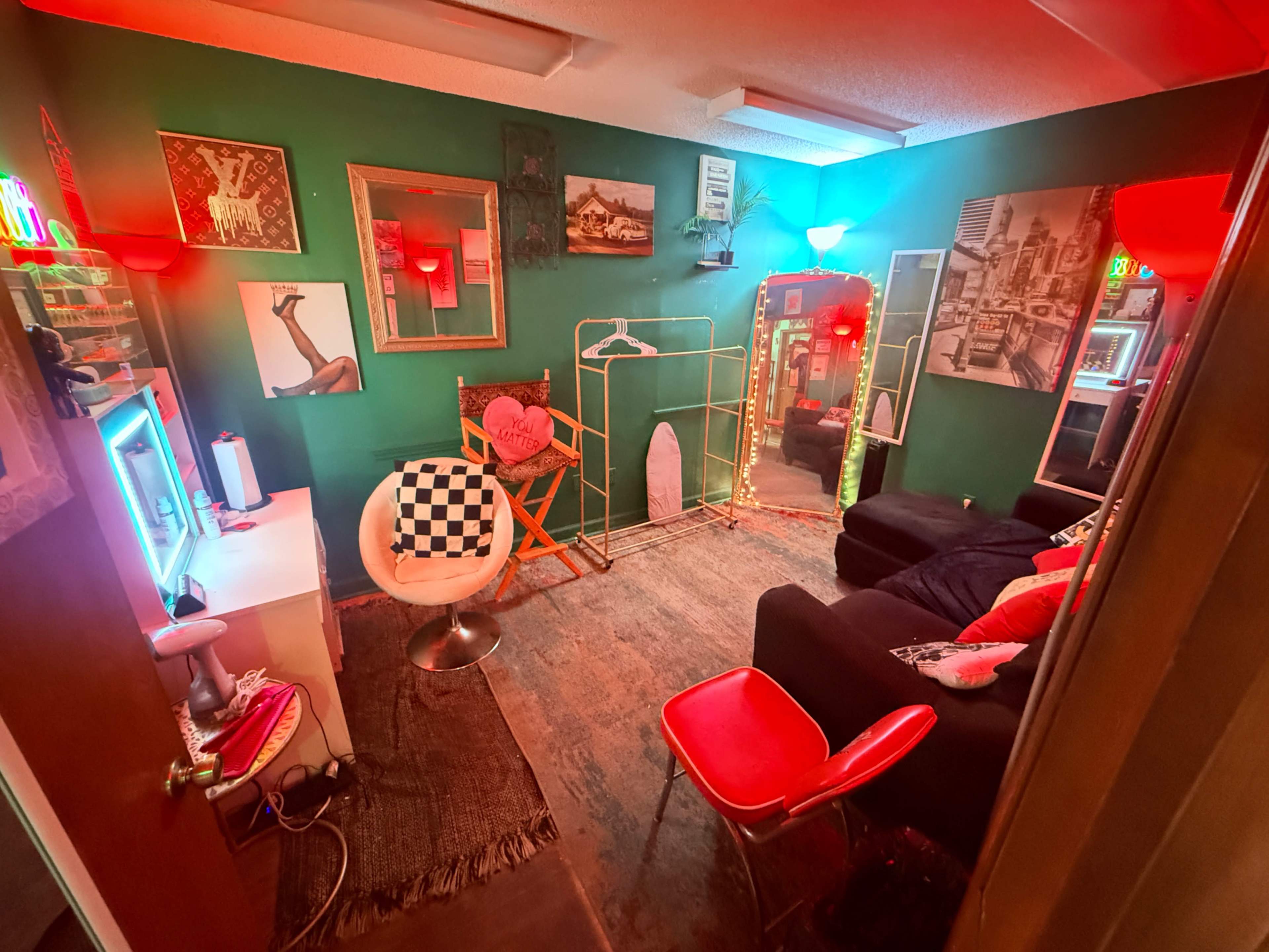 The image shows a cozy room with green walls, equipped with a black couch, a red chair, mirrors, and various decorative lighting and wall art.