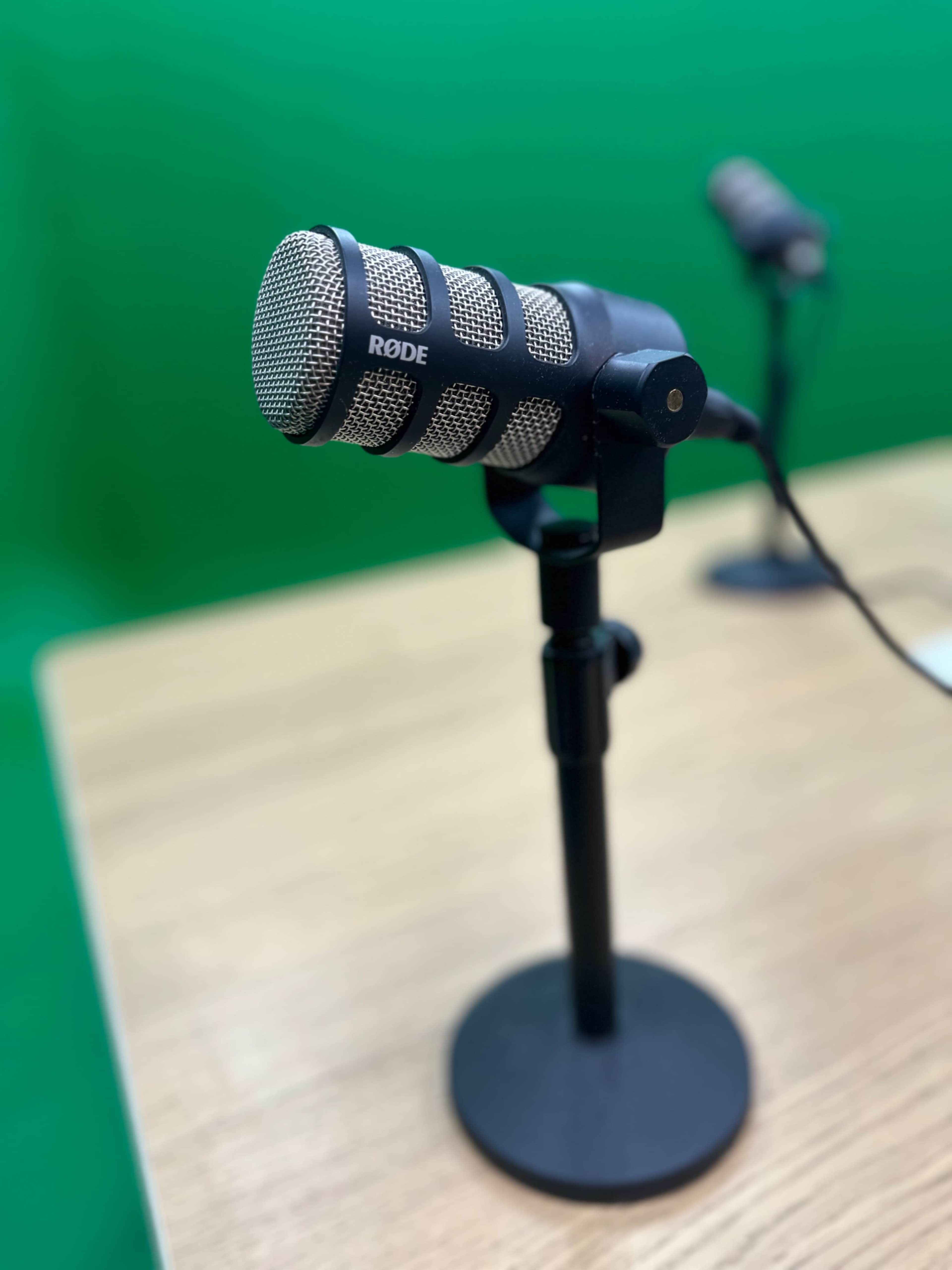 A Rode microphone is mounted on a stand on a wooden table, with a green backdrop in the background.