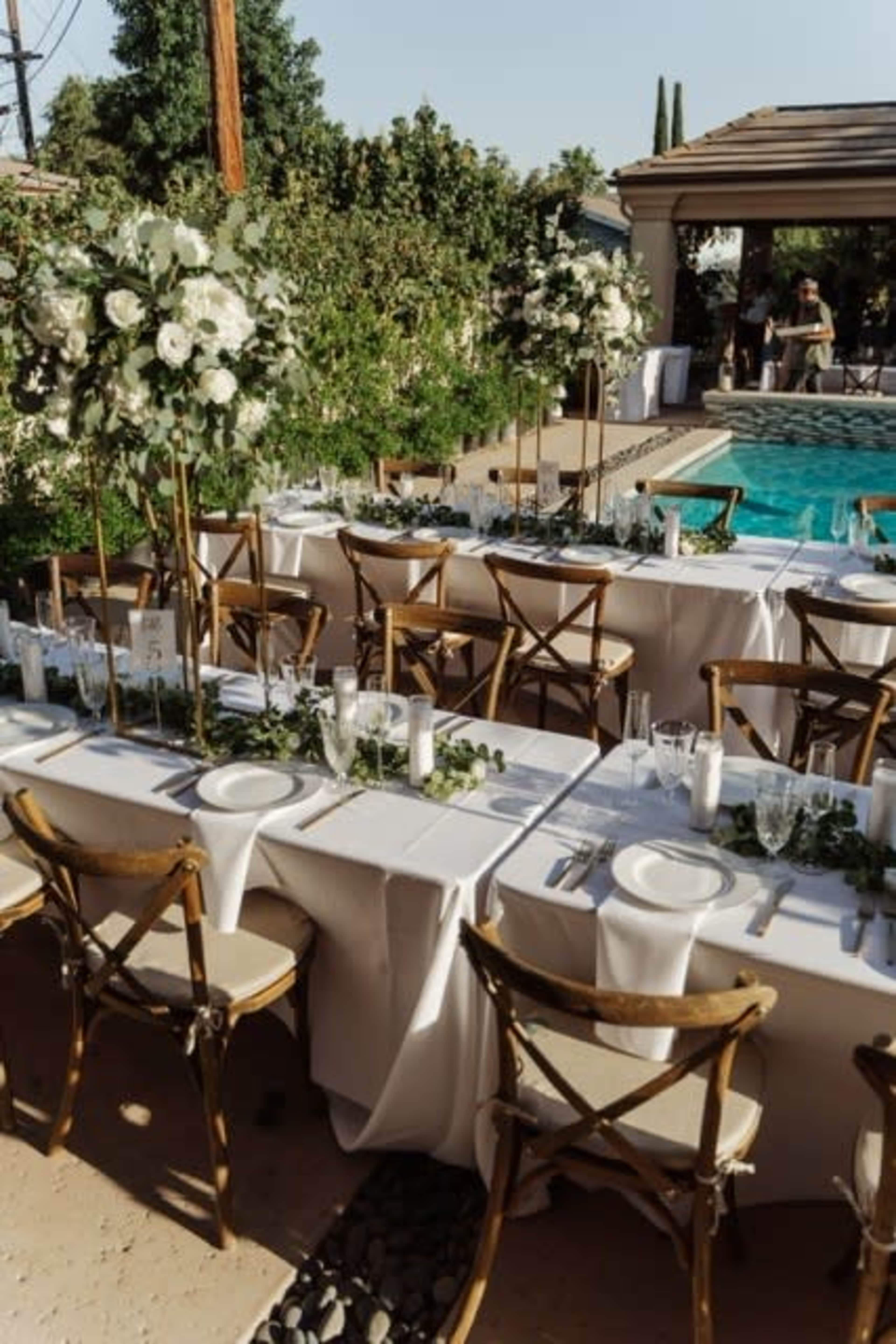 A long outdoor dining table is set near a pool, adorned with floral centerpieces and surrounded by brown wooden chairs.