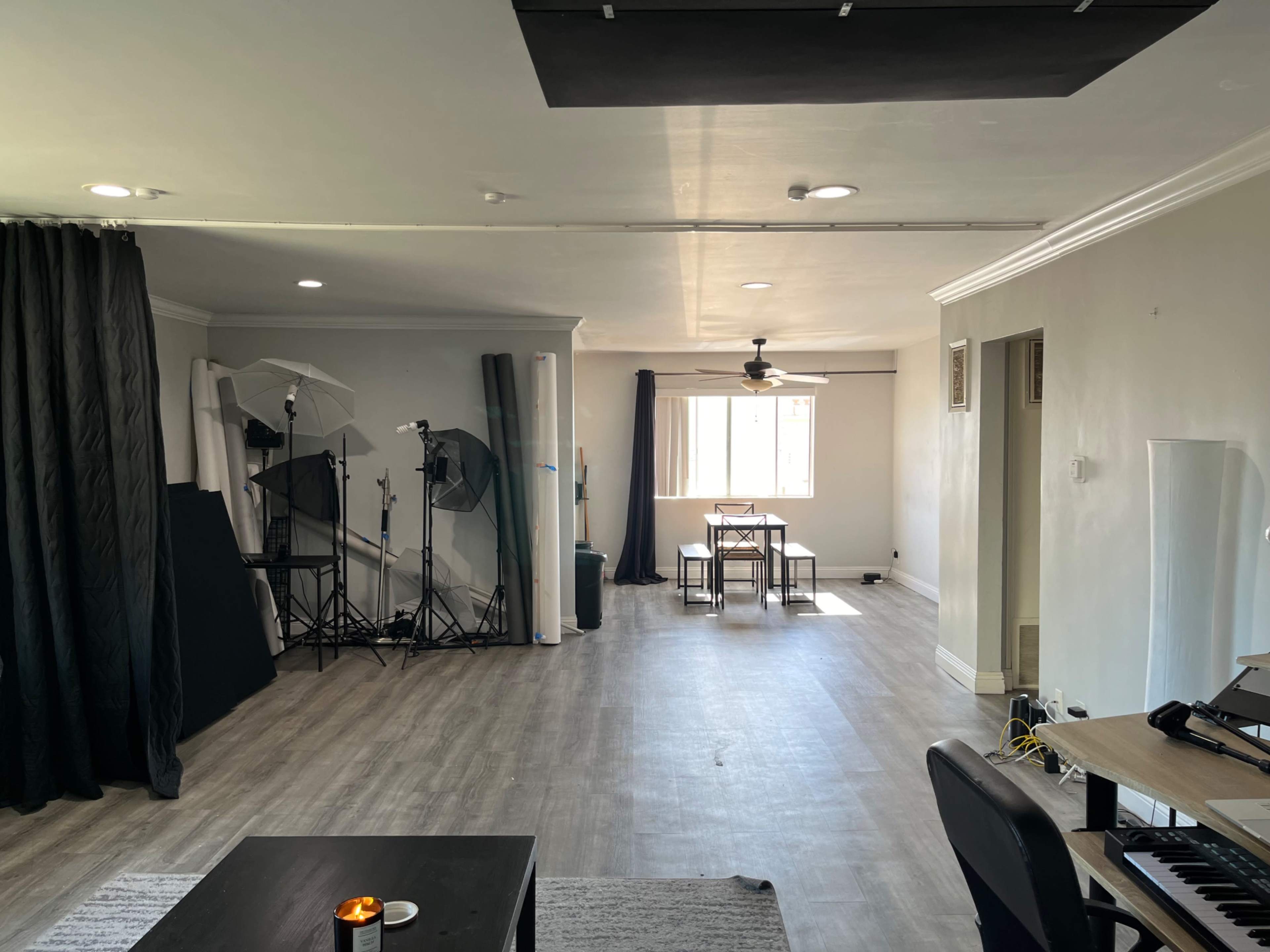 The image shows a spacious room with a light wood floor, featuring a small dining area with two tables, lighting equipment set up in one corner, and a workspace with a keyboard along one wall.