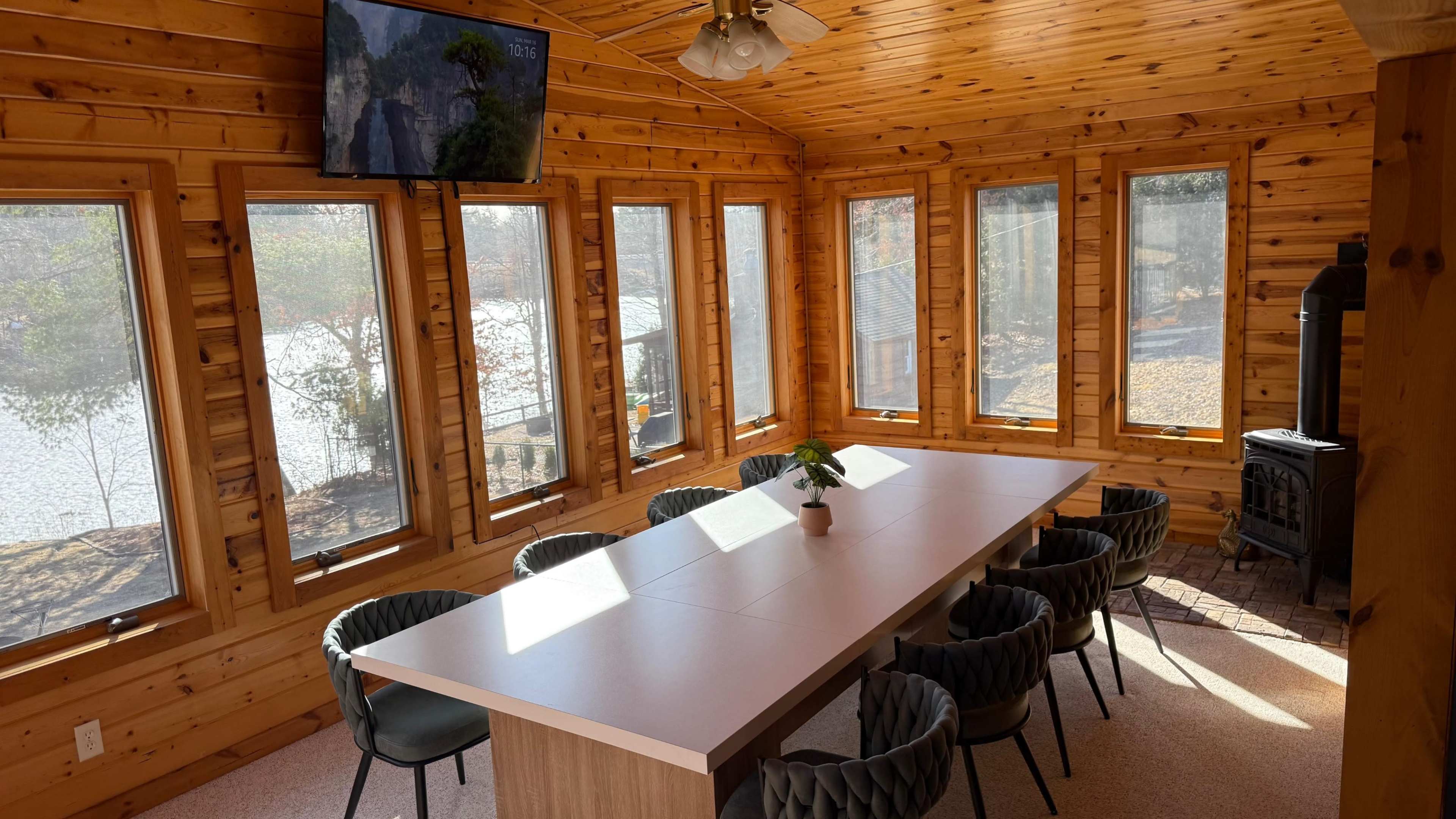 The image shows a wooden sunroom with large windows, a long white table, and several black chairs arranged around it.