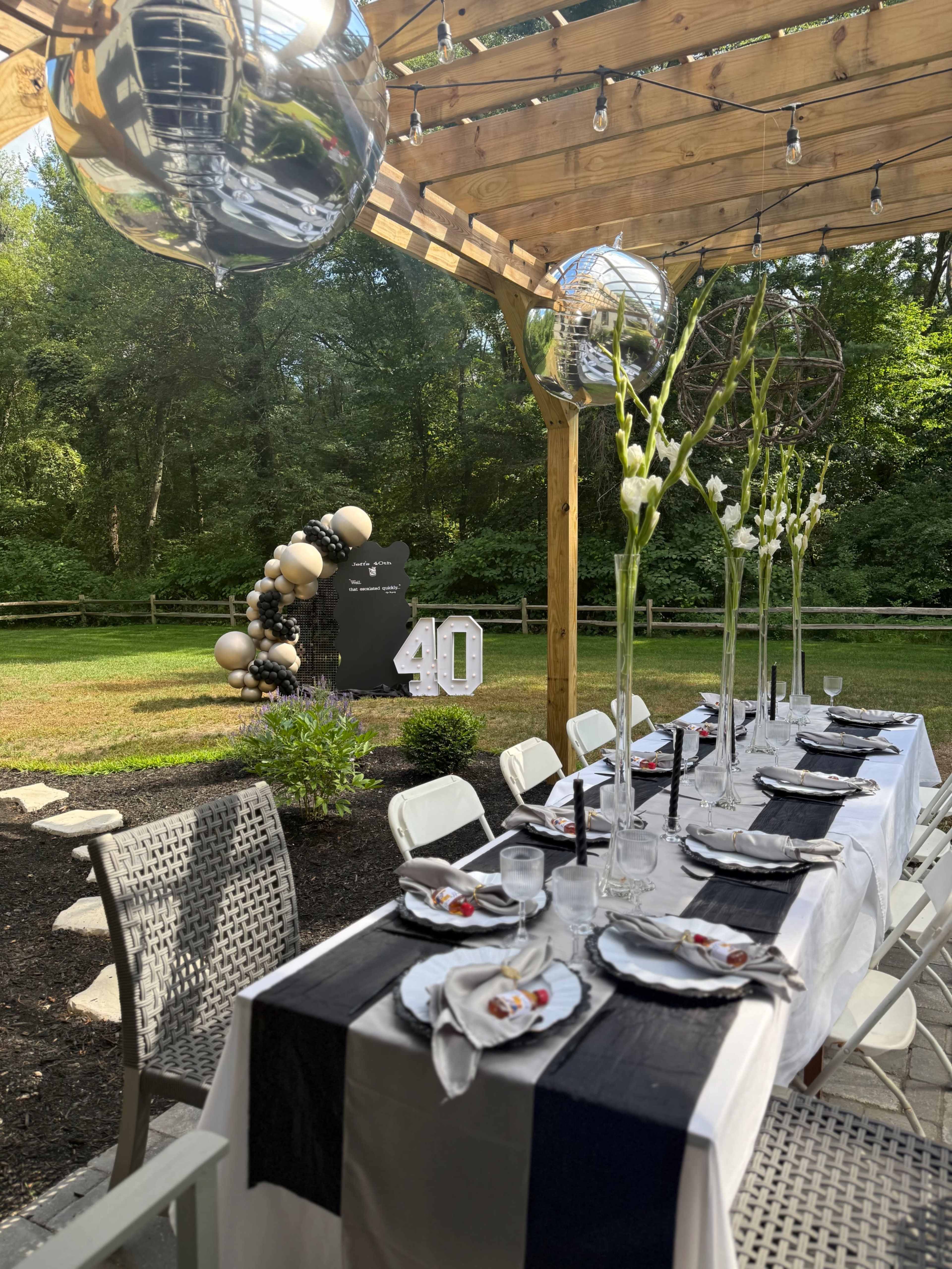 A long dining table is set under a wooden pergola, adorned with black and white table settings, and is decorated for a 40th birthday celebration in a green backyard.