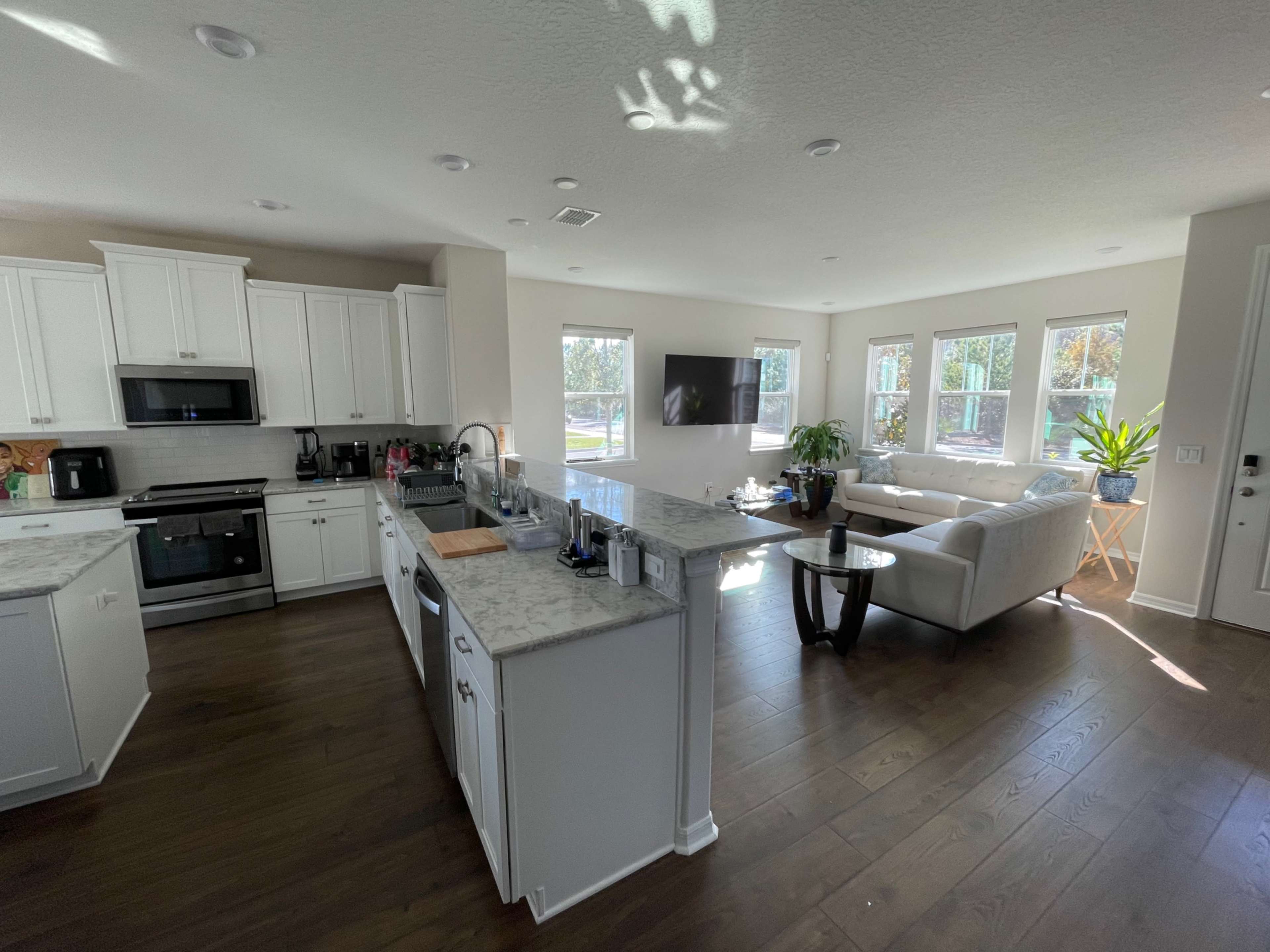 A modern open-concept living space features a kitchen with white cabinets and a marble countertop adjacent to a seating area with a sofa and large windows.