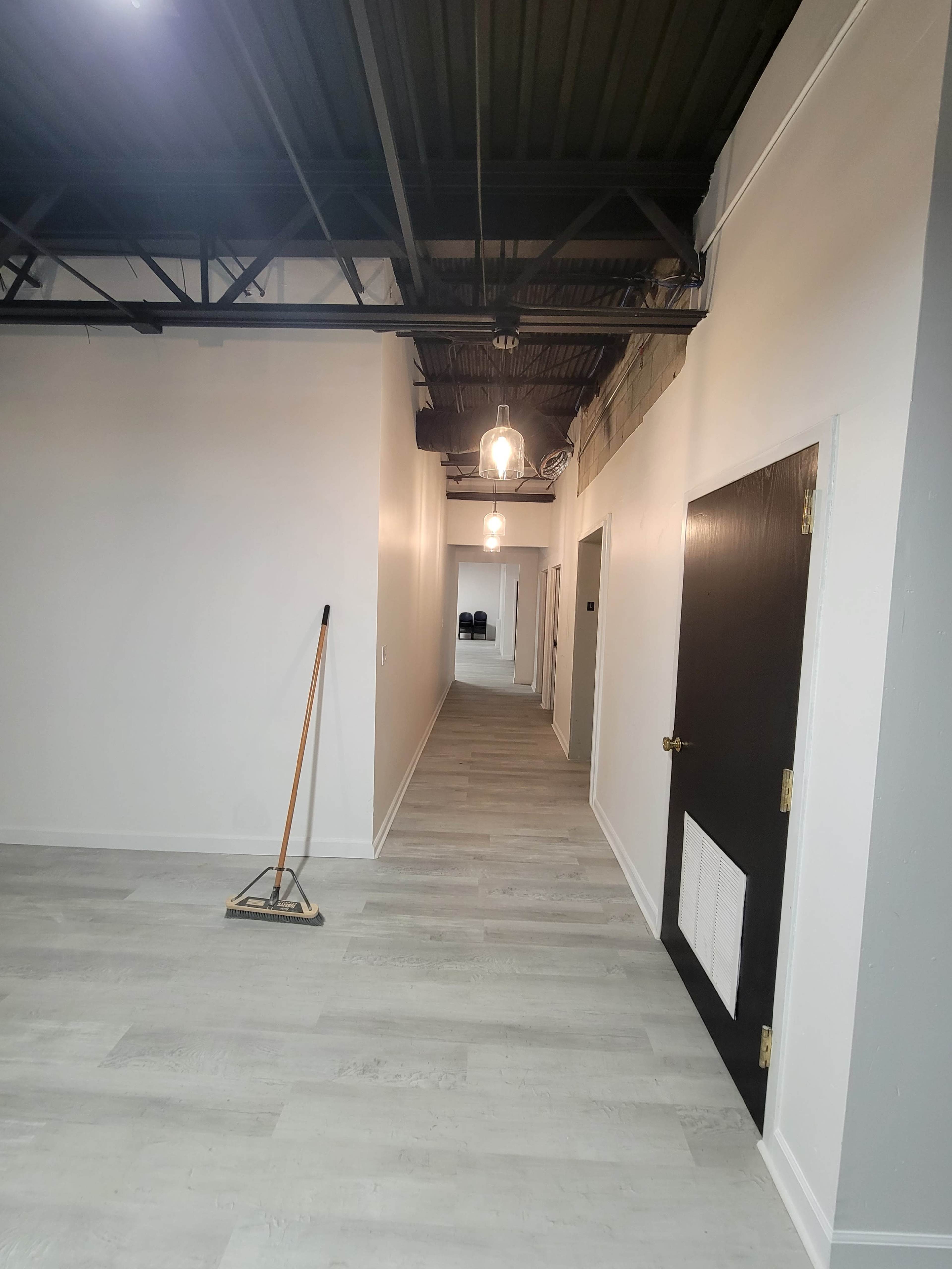 A long, empty hallway with wooden flooring, white walls, and overhead lighting leads to a closed door at the far end.