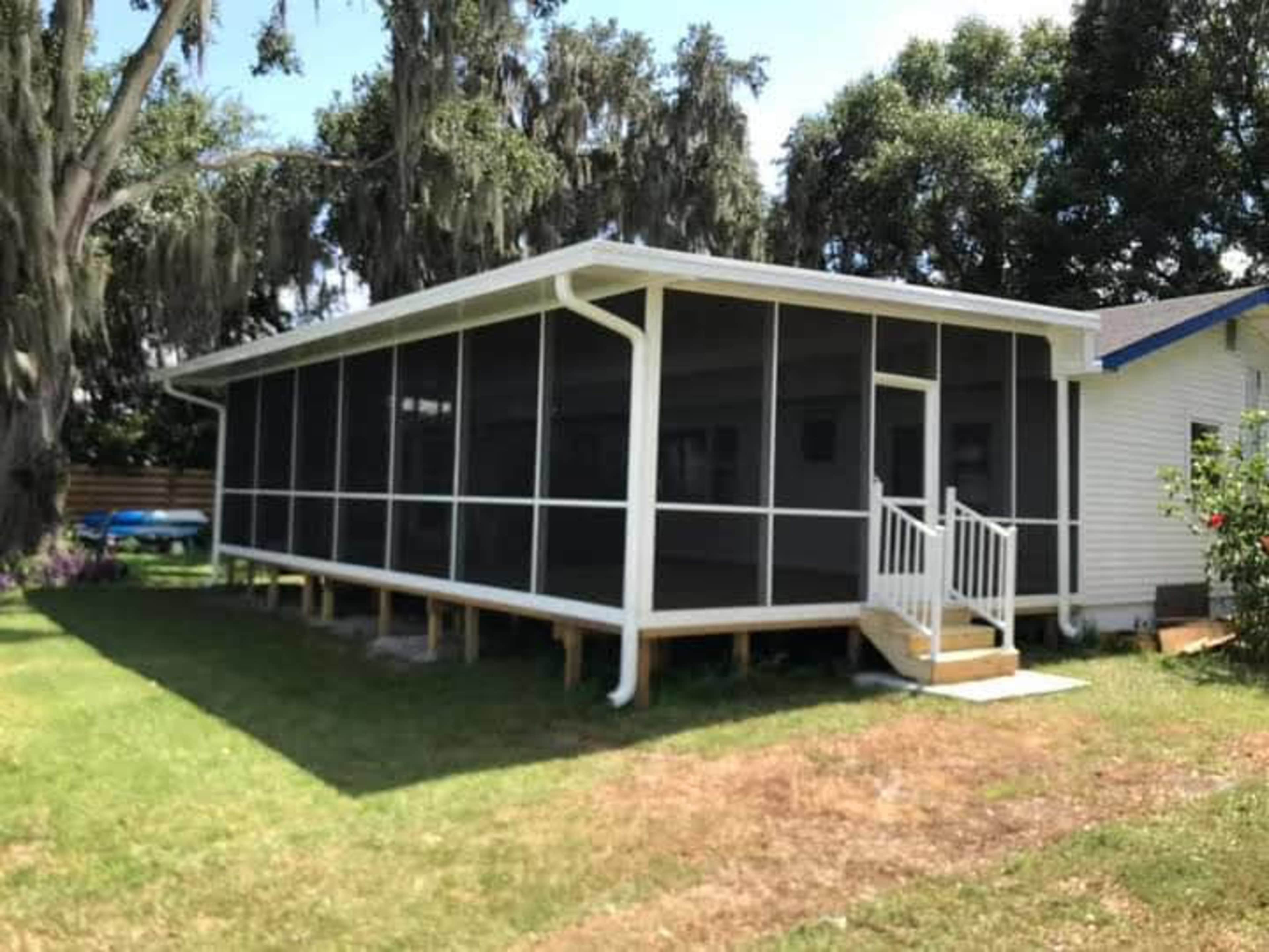 The image shows a large screened porch attached to a house, situated on a grassy area with trees in the background.