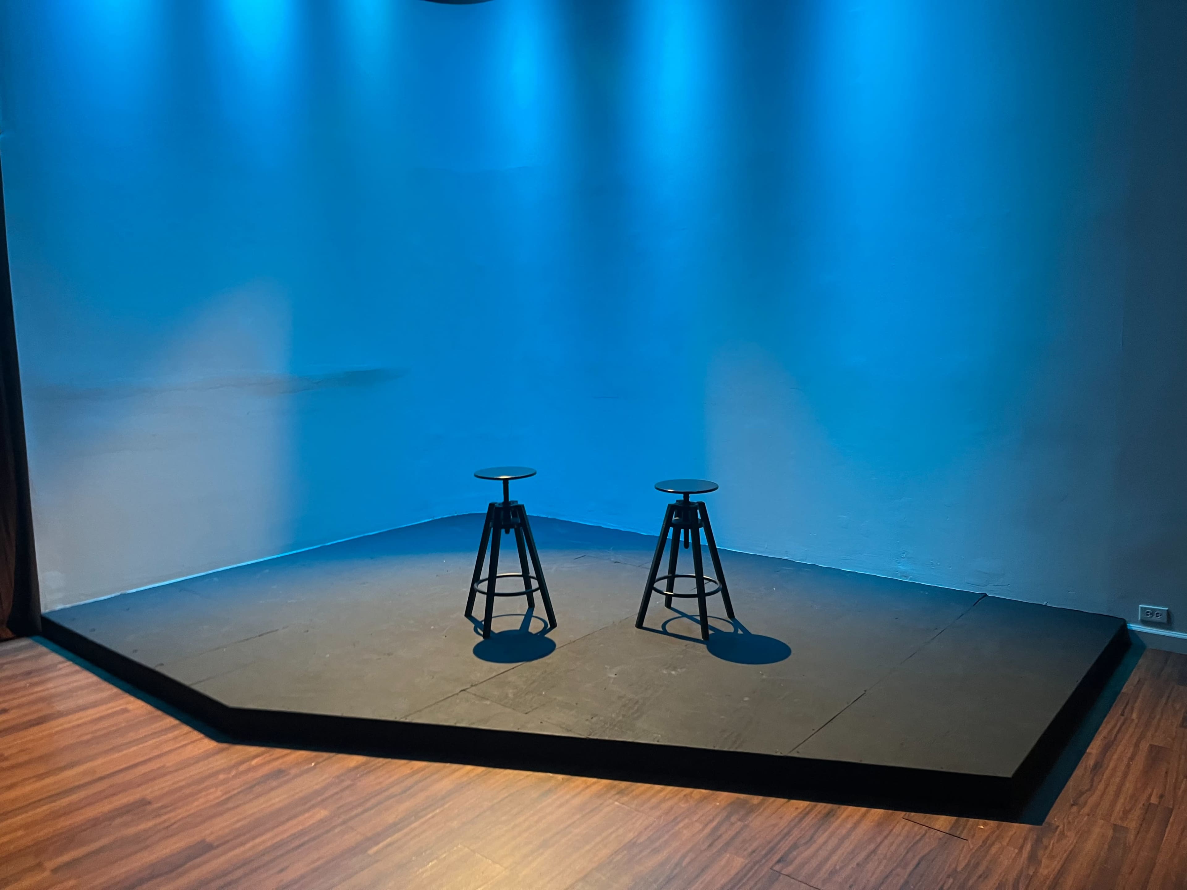 The image shows a small stage illuminated by blue lighting, featuring two black stools positioned on a wooden floor.
