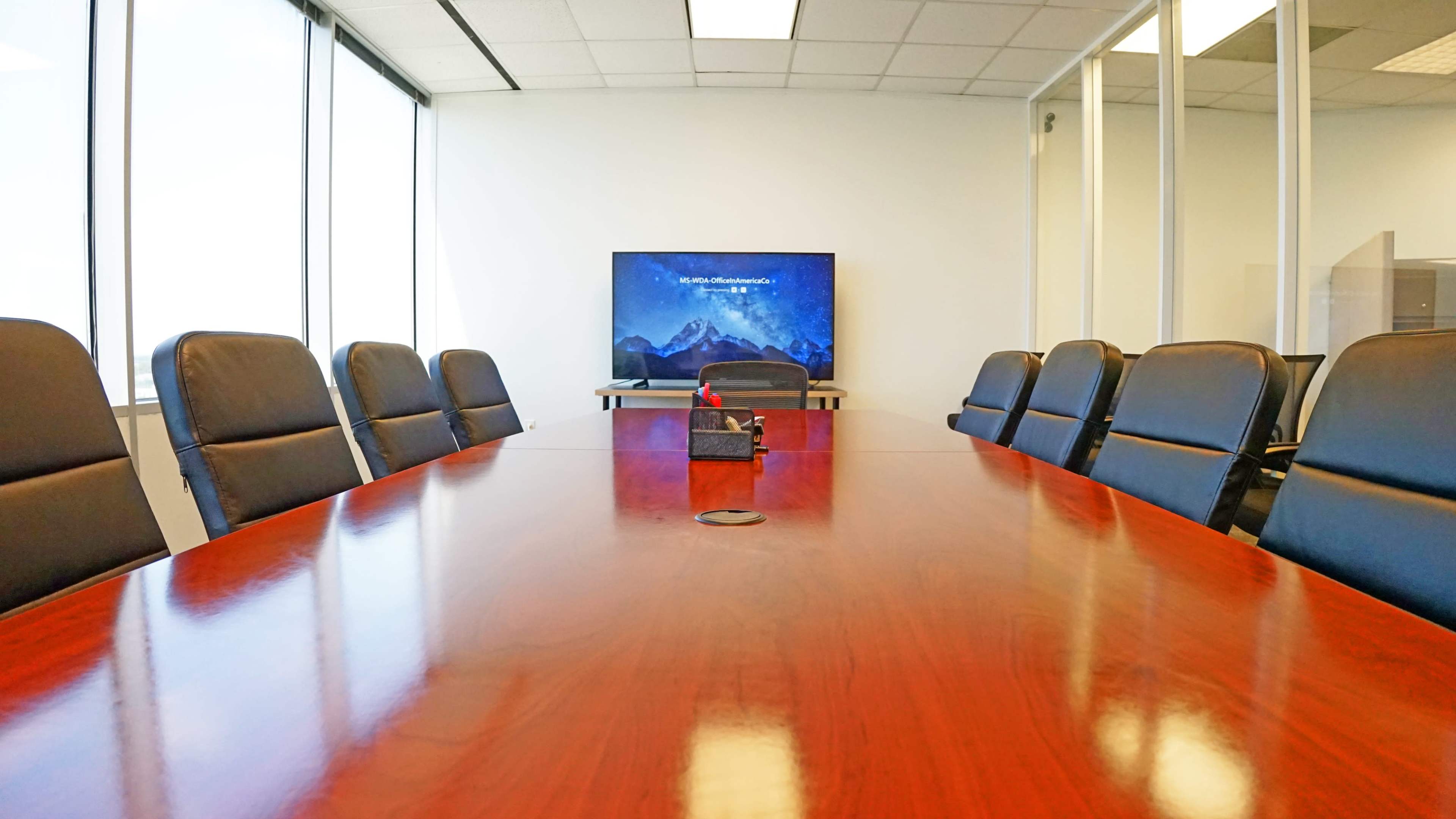 A long wooden conference table is set with black leather chairs in a well-lit meeting room featuring a window and a screen displaying a mountain scene.