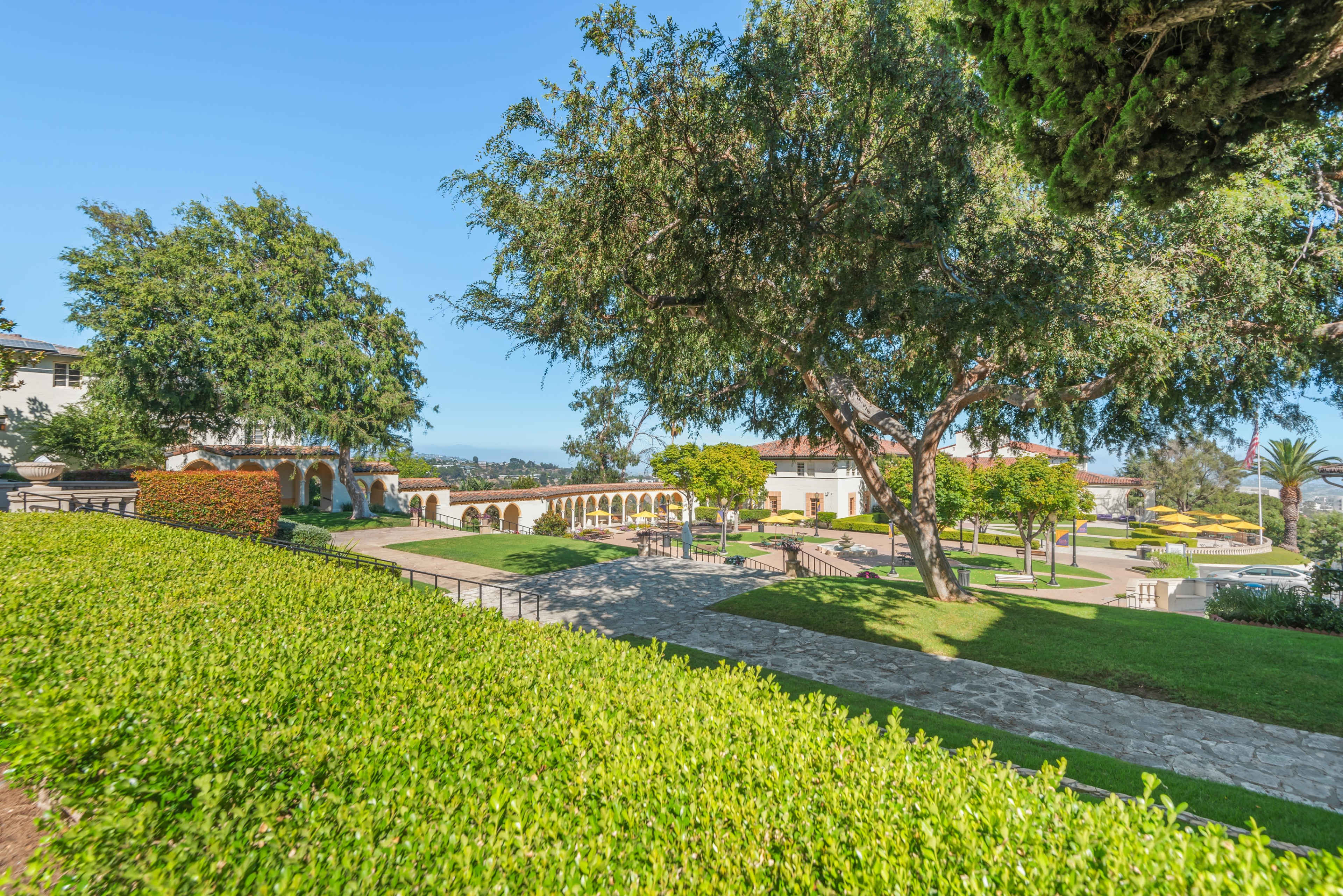 The image shows a landscaped garden area with trees, hedges, and pathways, leading to a building in the background under a clear blue sky.
