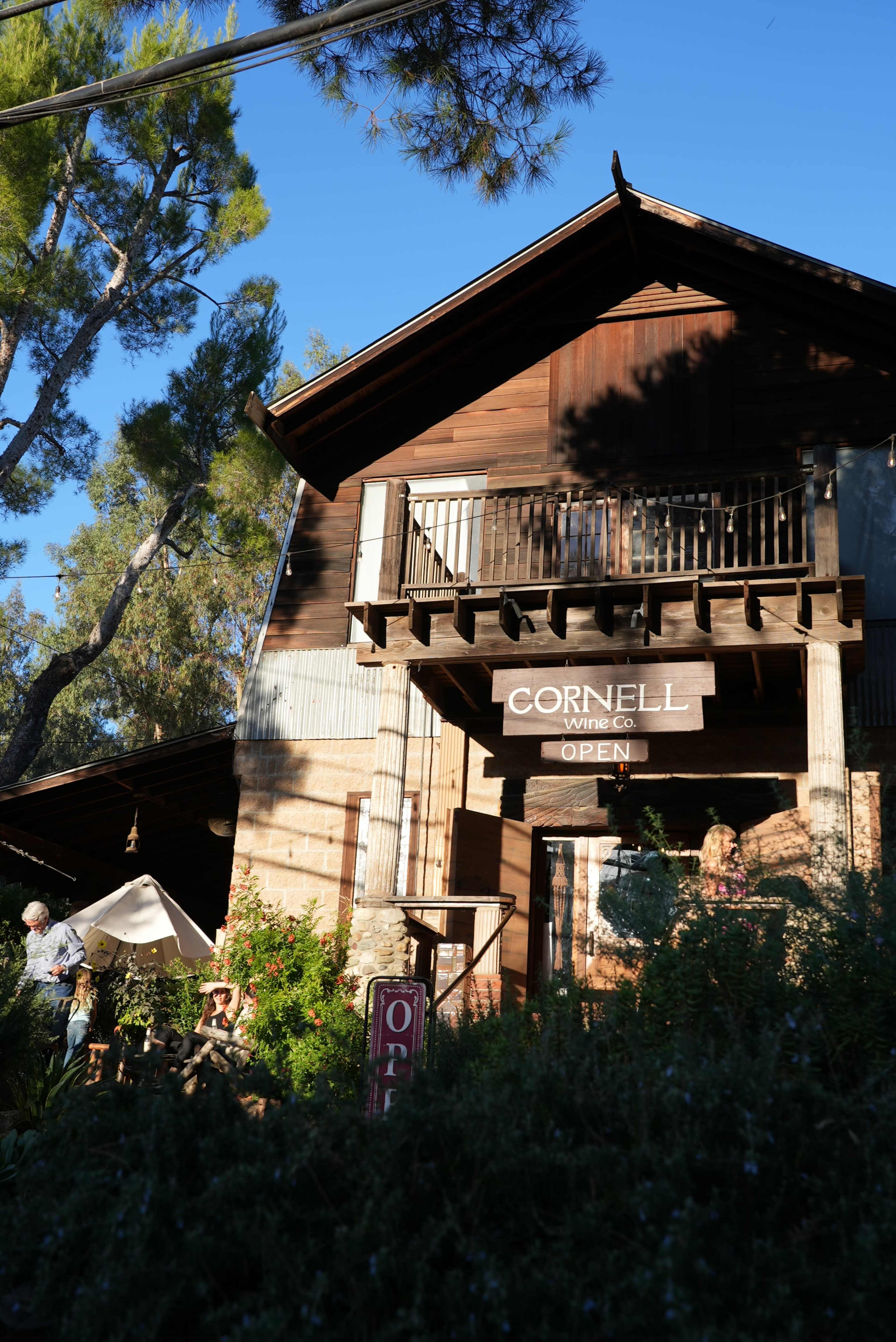 The image shows a wooden building labeled "CORNELL" with an "OPEN" sign, surrounded by trees and greenery.