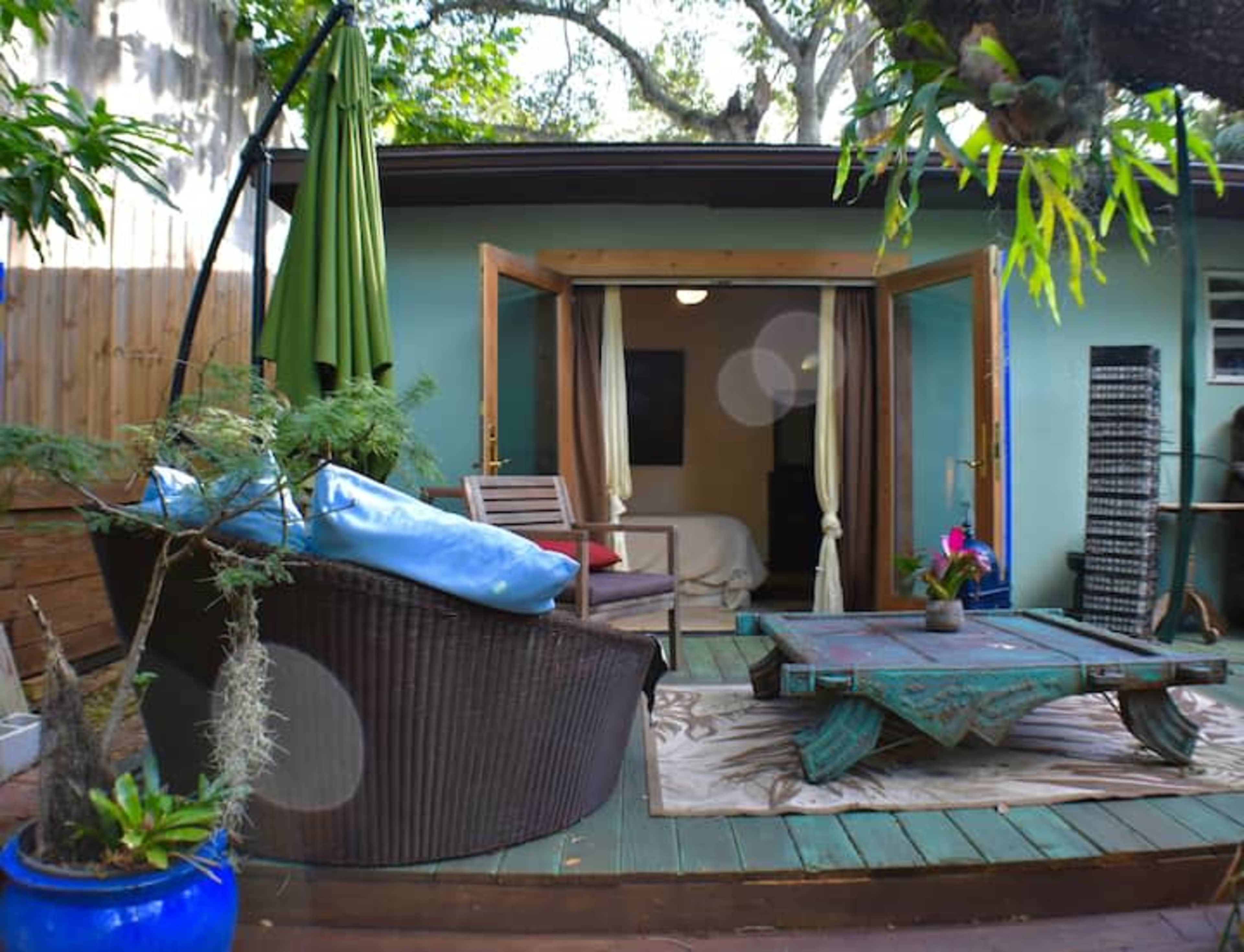 A cozy outdoor seating area features a wicker chair, a small table, and open doors leading into a room with a light green wall.