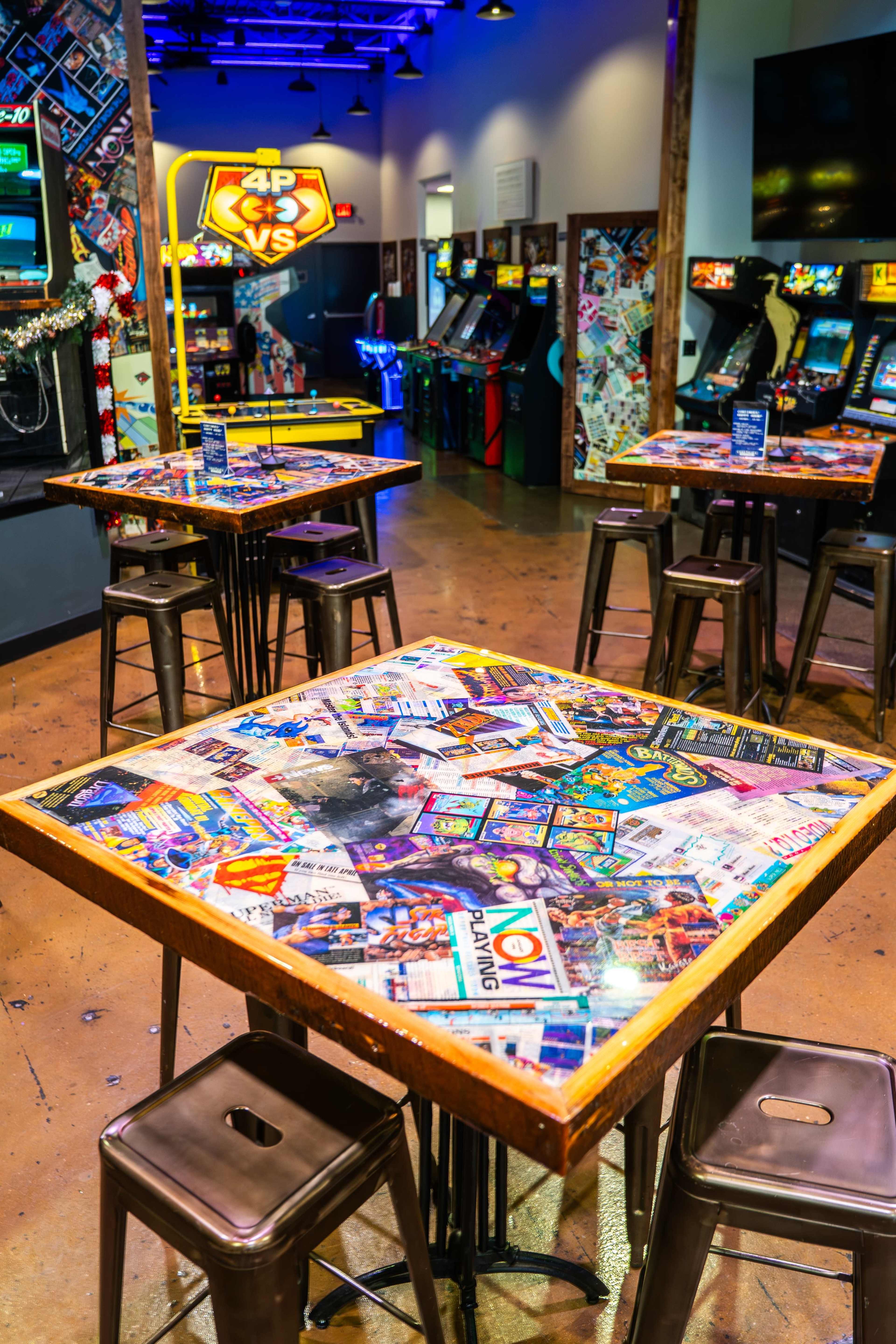 A vibrant arcade room features several metal tables adorned with colorful game-related memorabilia, surrounded by vintage arcade cabinets.