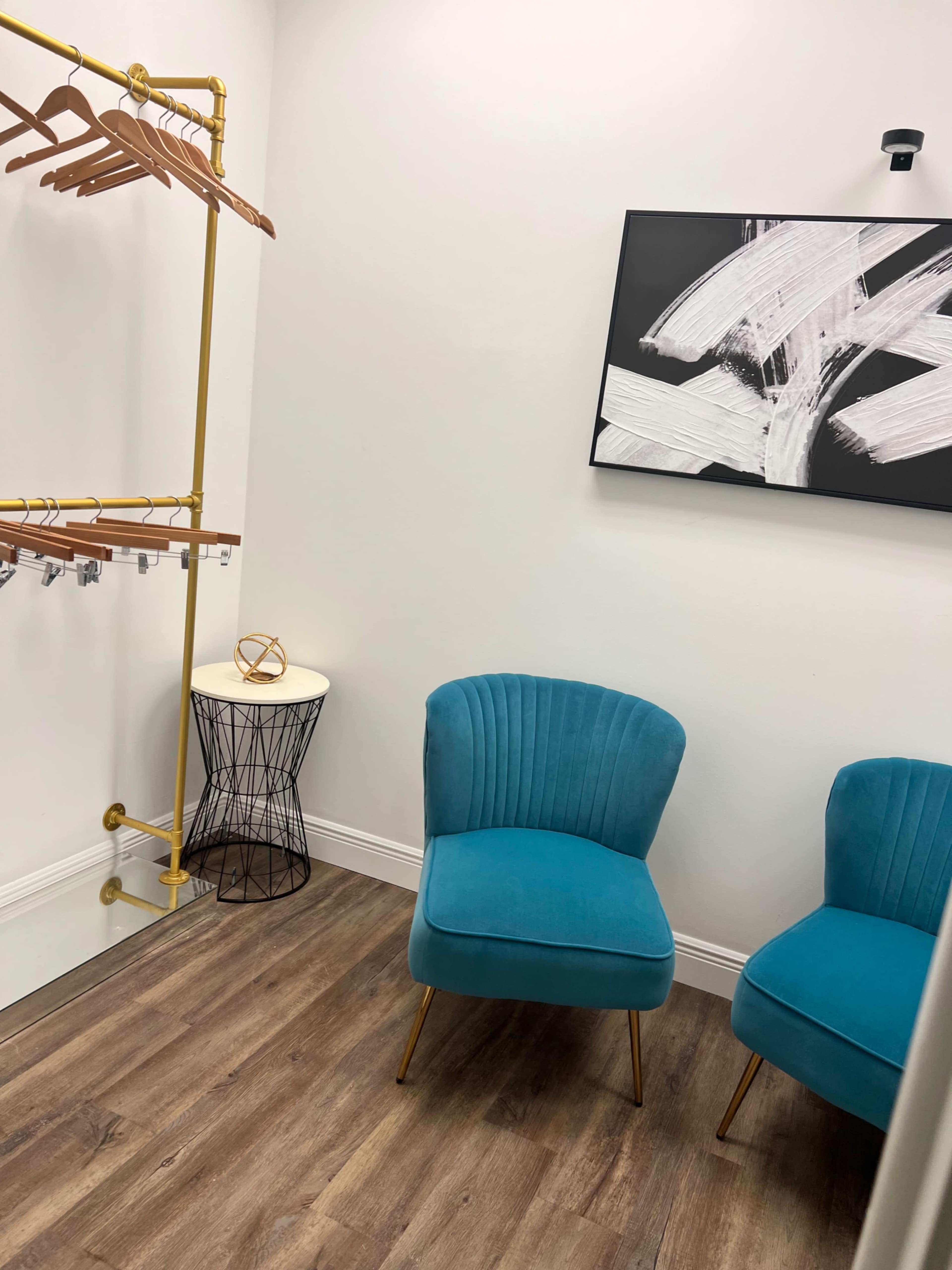The image shows a small room with two turquoise chairs, a clothing rack, a black wire side table, and a wall art piece.
